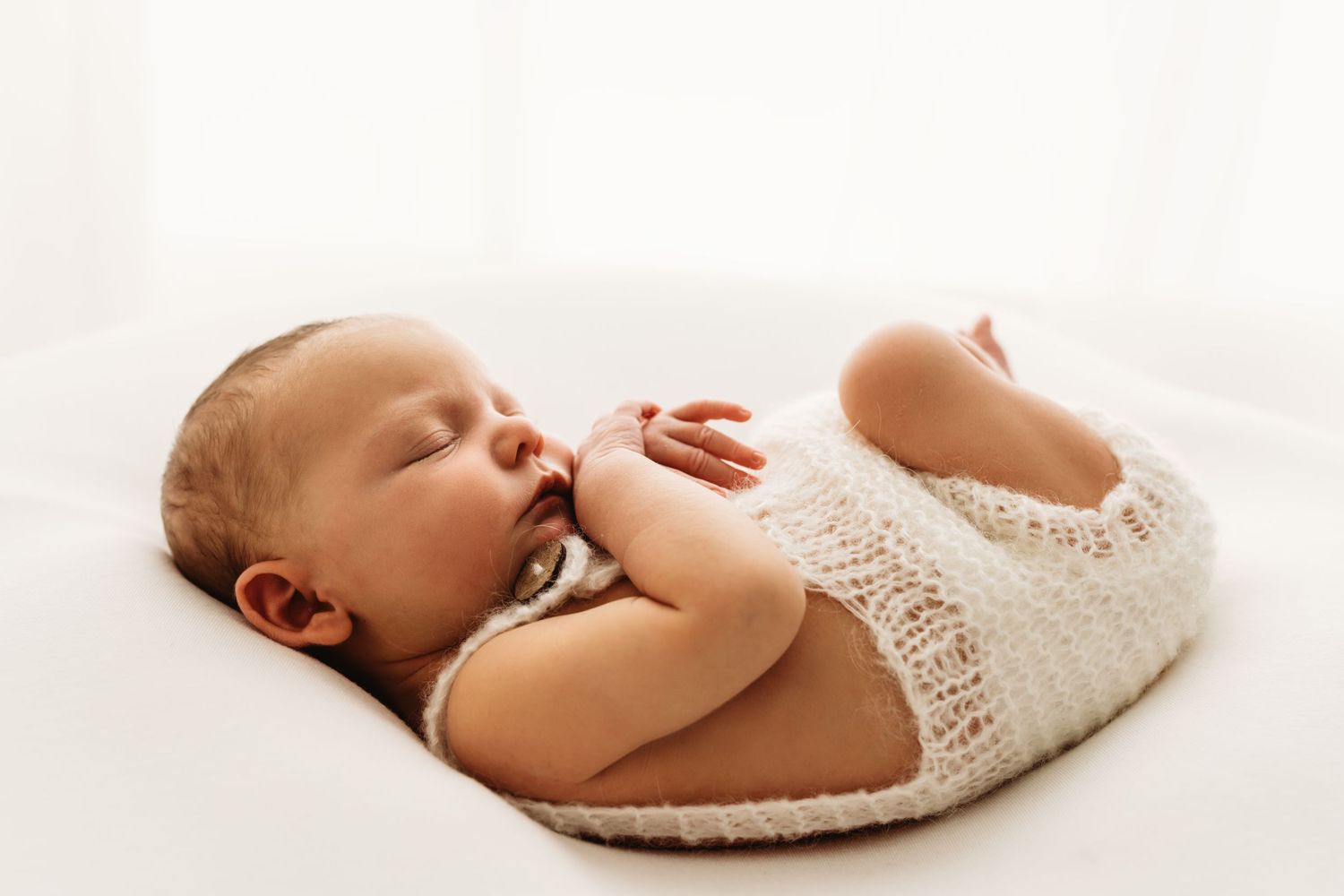 Peaceful newborn baby sleeping curled up in a cream knit wrap on a white background during a newborn photography session