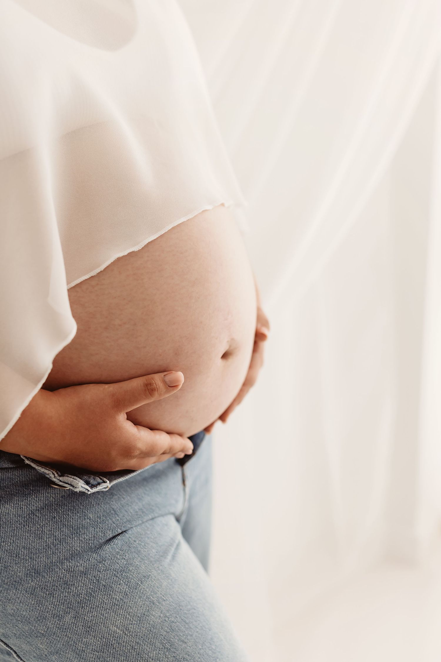 Close-up of pregnant woman cradling baby bump, wearing white blouse and blue jeans near sheer curtains.