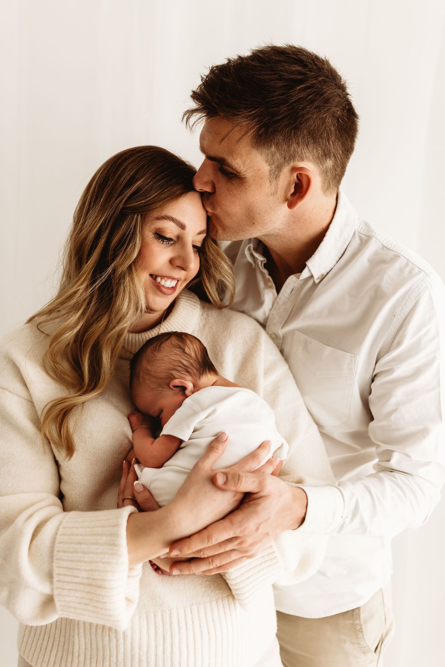 Happy couple in white sweaters tenderly holding their sleeping newborn baby in a bright, airy studio setting.