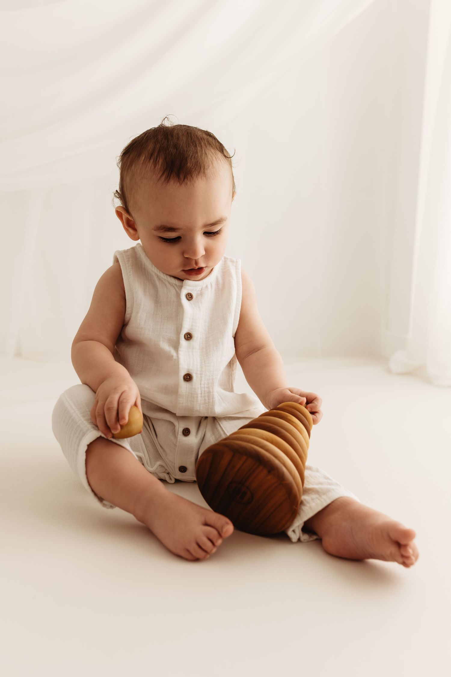 Baby in white linen romper sitting on floor playing with wooden stacking ring toy.
