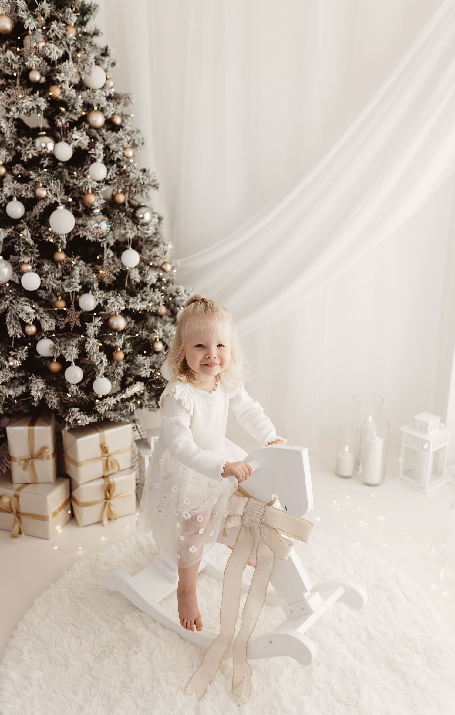 Smiling toddler girl in white dress on rocking horse beside decorated Christmas tree with gifts.
