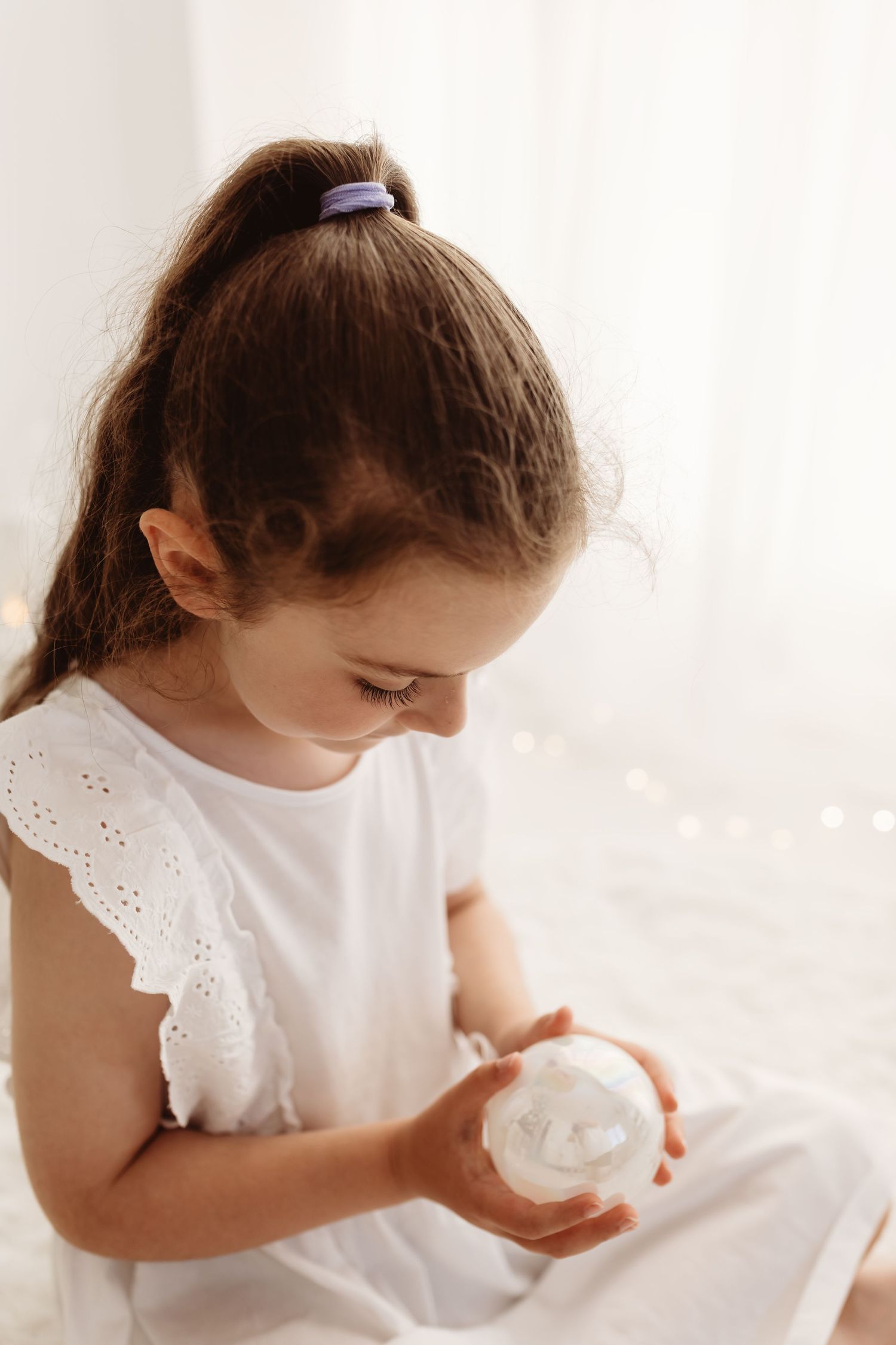 Young girl in white dress holding a snow globe, with bokeh lights in the background.