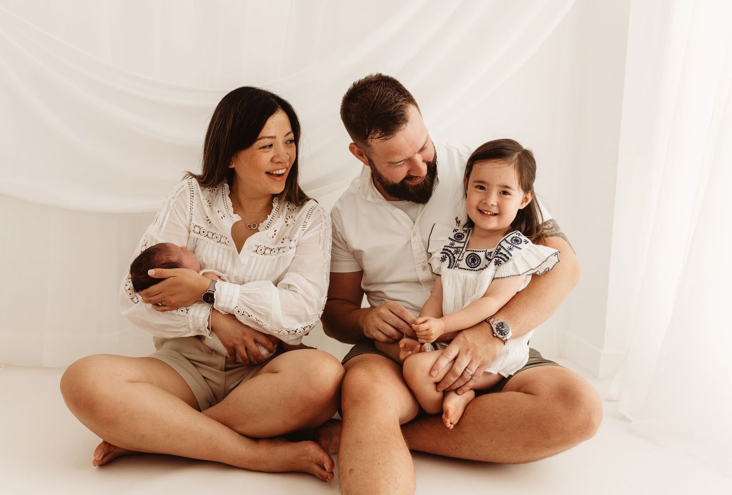 Happy family of four sitting together, parents holding newborn baby and toddler girl in white studio setting.