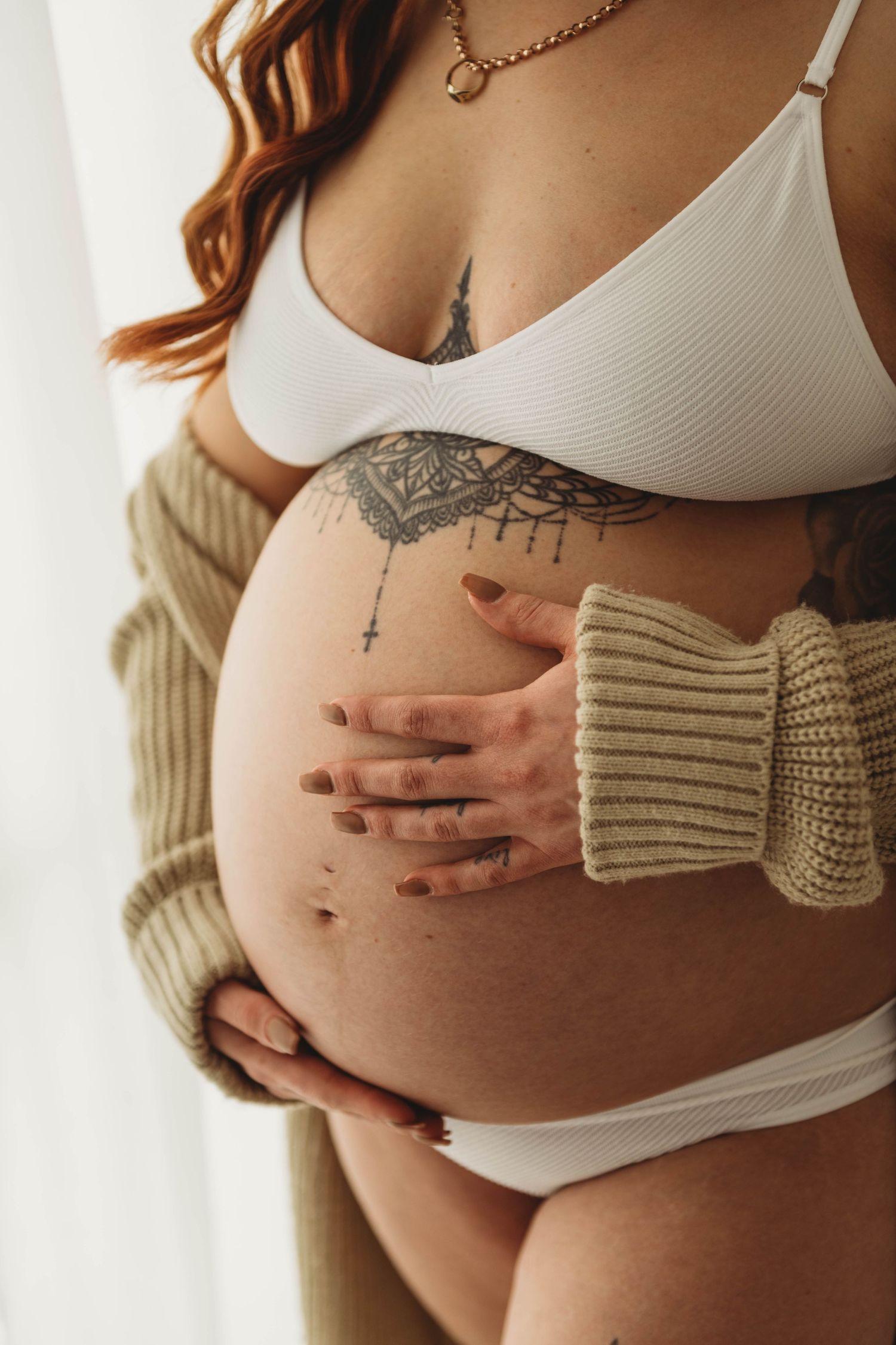 Close-up of a pregnant belly with mandala tattoo and hands in white crop top.