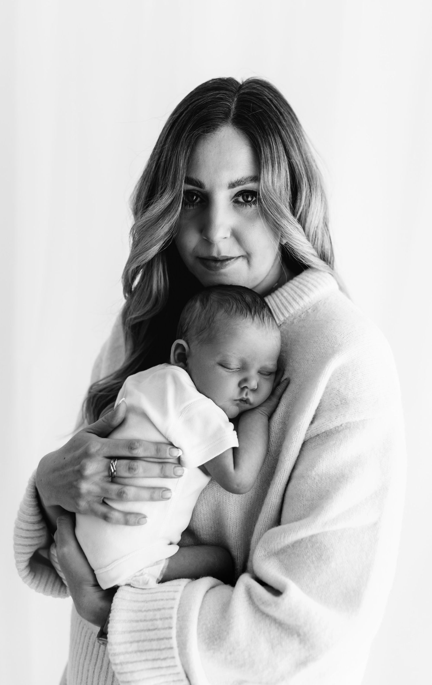 Black and white photo of a woman in a cozy sweater tenderly holding a sleeping newborn baby wrapped in white cloth.