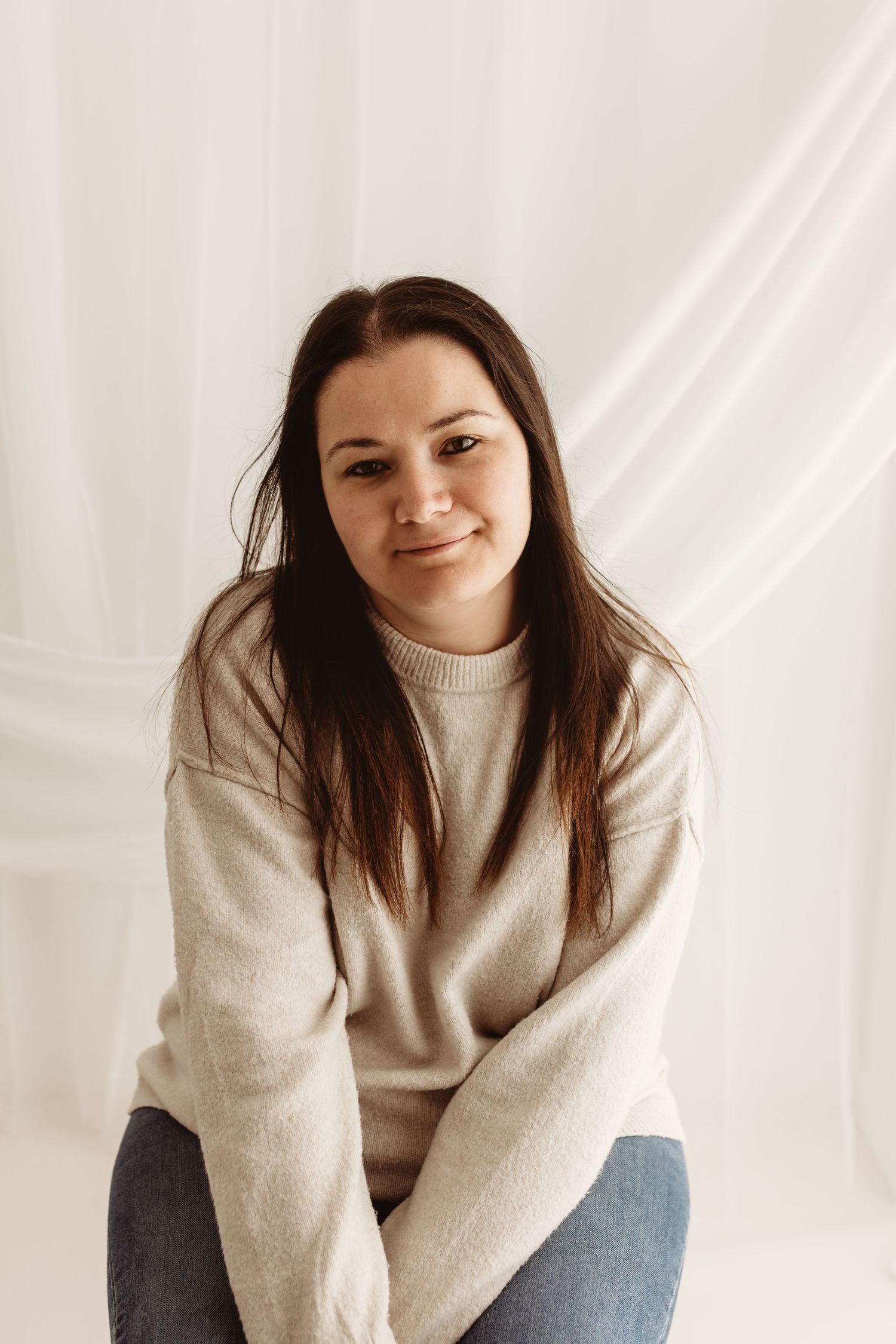 Young woman with dark hair smiling, wearing a beige sweater and jeans, seated against a white fabric background.