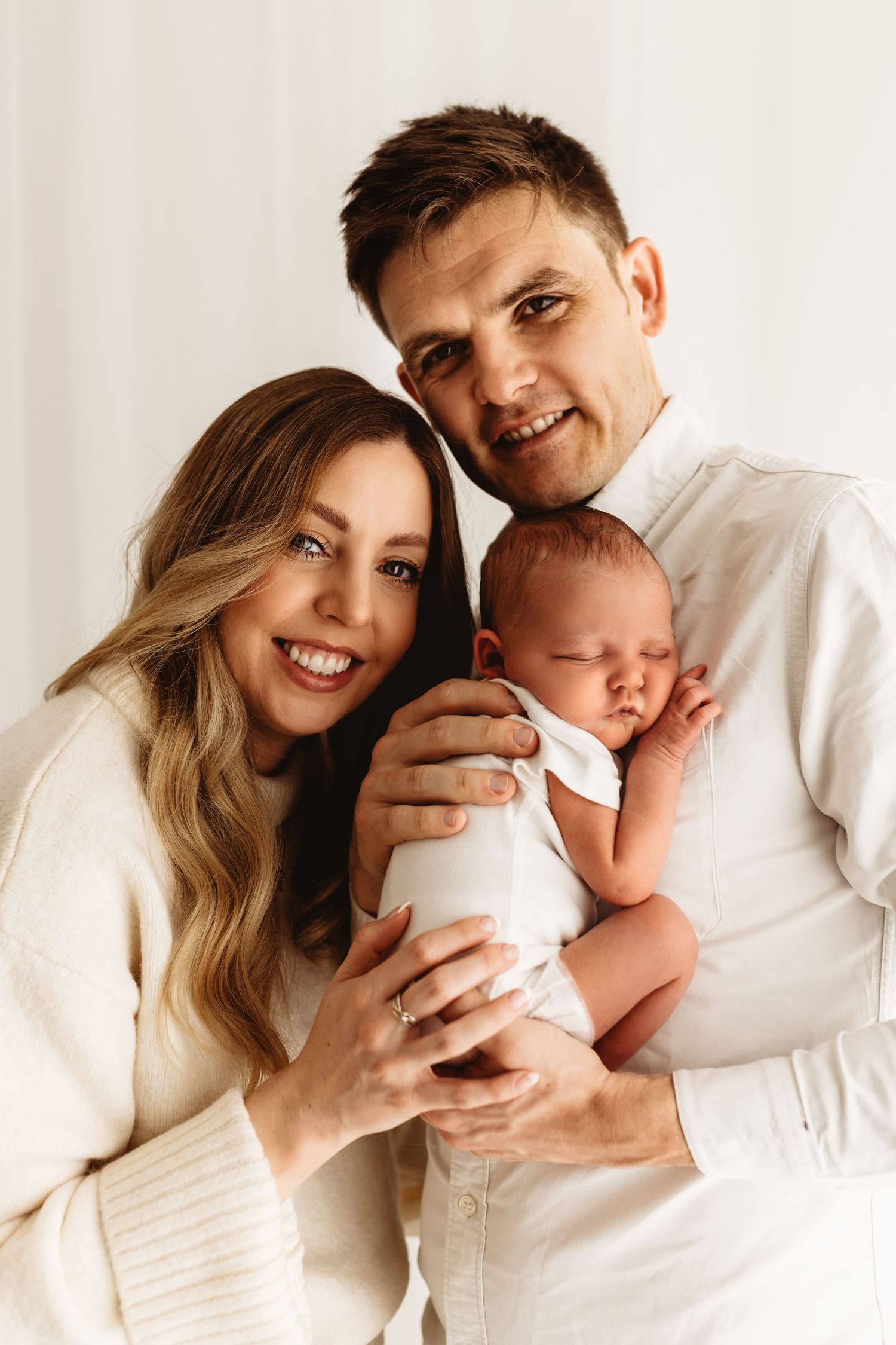 Happy young parents smiling and holding their sleeping newborn baby, dressed in white, against a light background.
