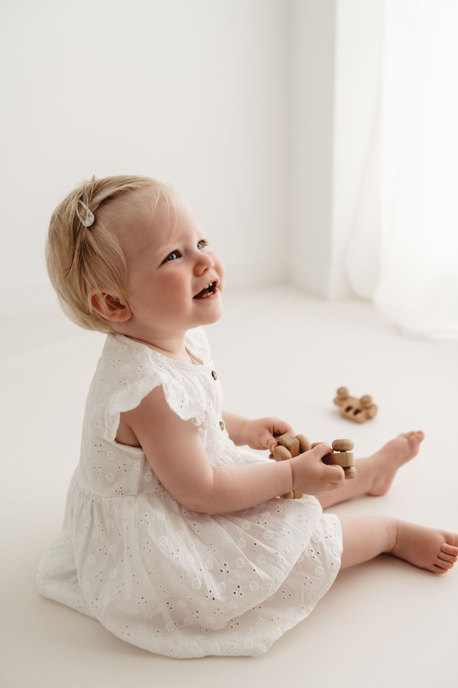 Baby in white eyelet dress sits on white floor with wooden toys during a Sitter photography session in Corby, Northamptonshire