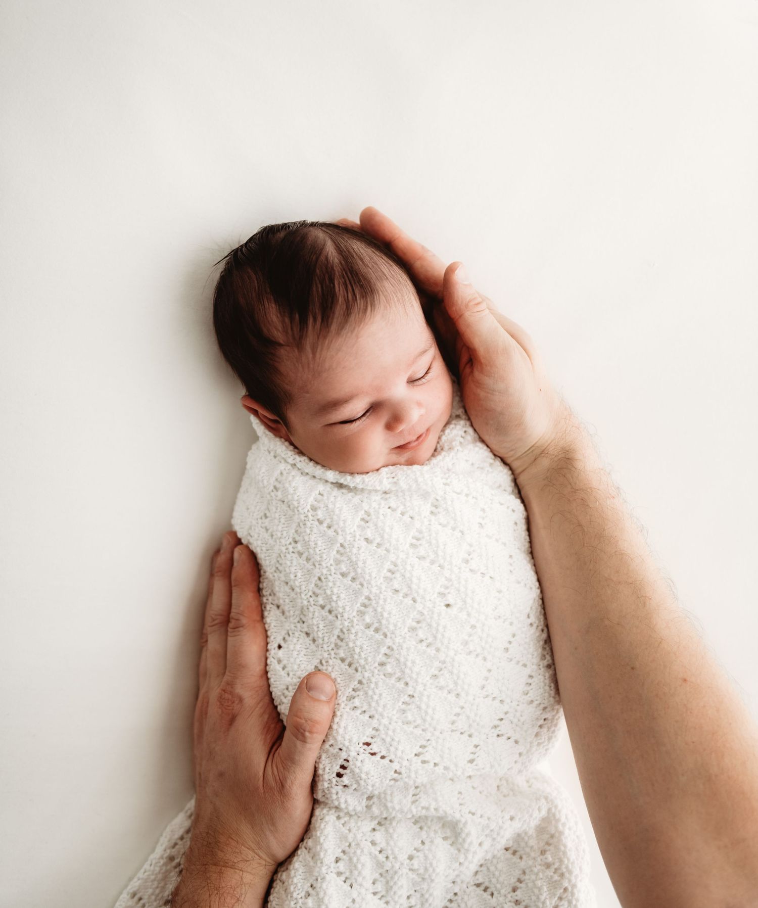 A newborn baby swaddled in white lace fabric is gently held by adult hands against a light background.