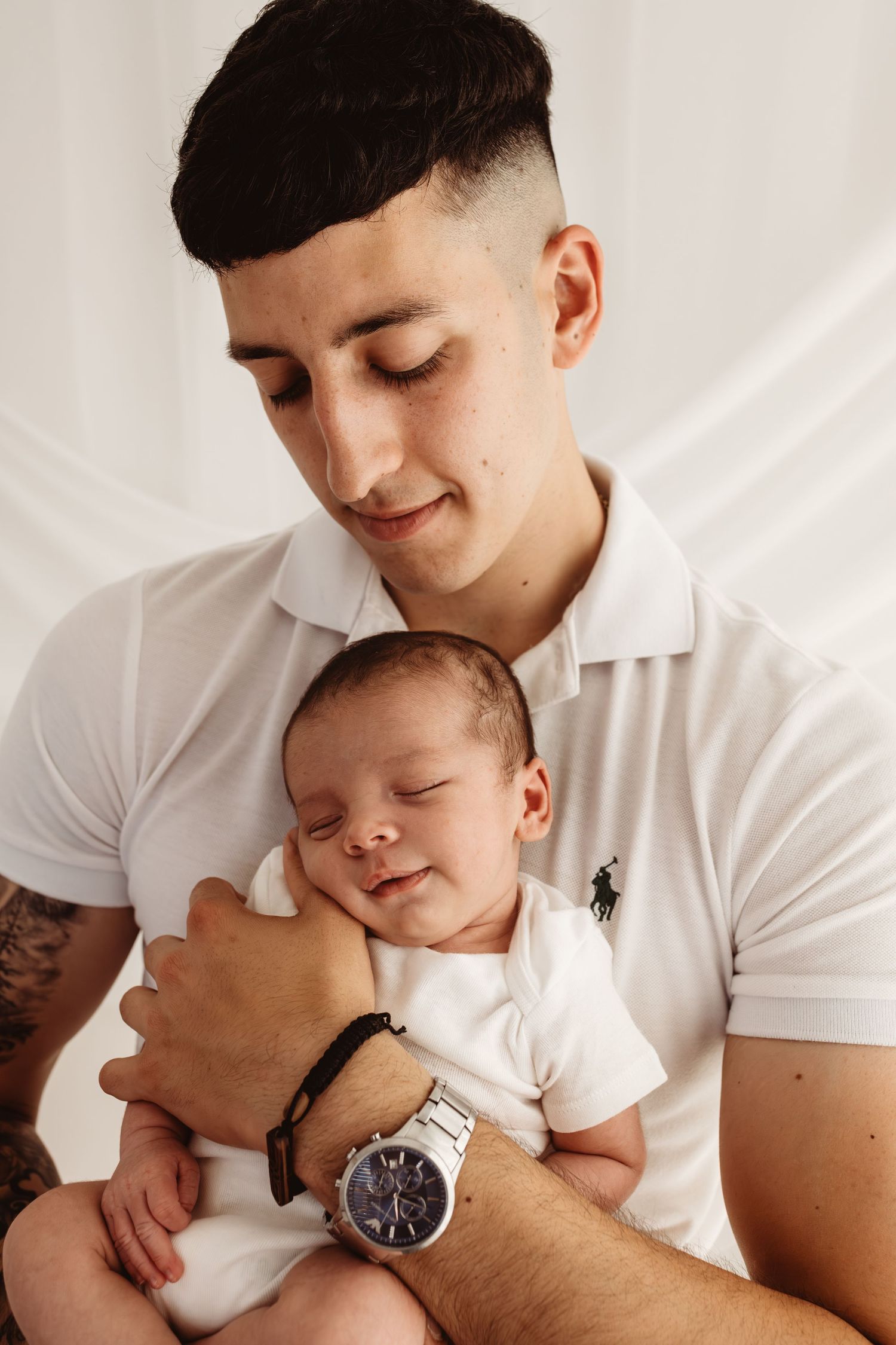 Young father in white polo shirt tenderly holds sleeping newborn baby, wearing a silver watch.