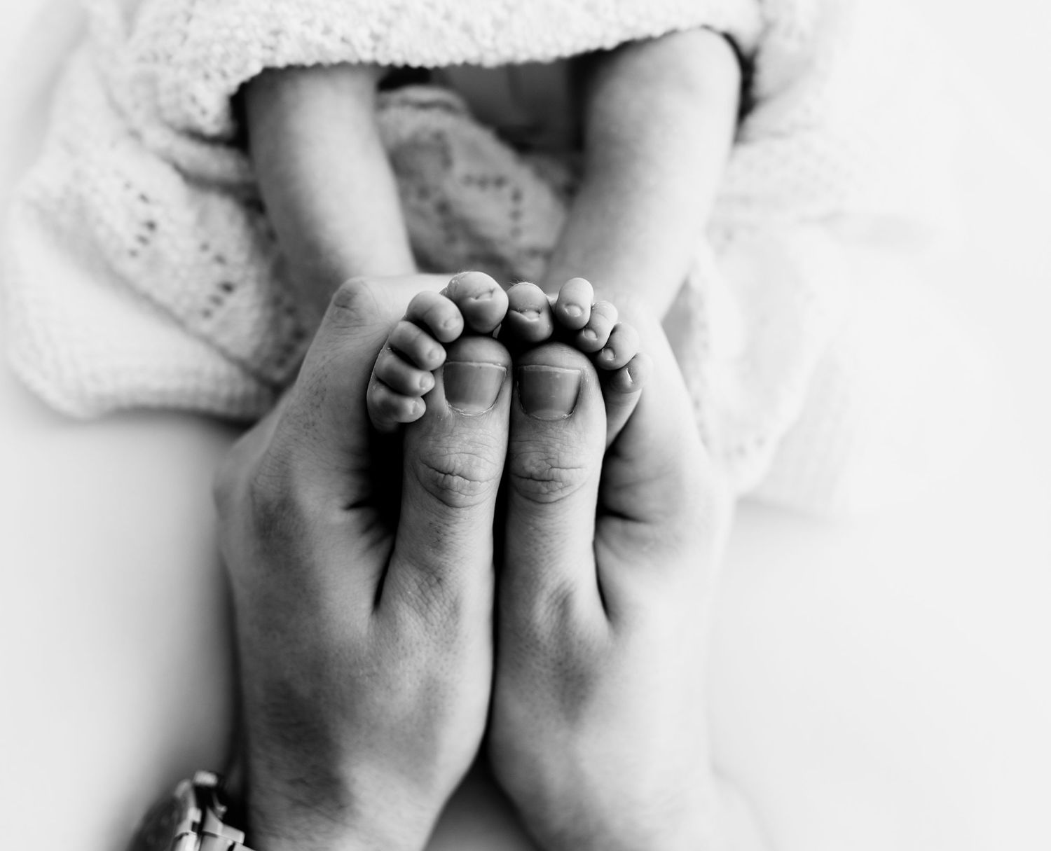 Black and white photo of adult hands gently cradling tiny newborn baby feet, wrapped in a soft white blanket.