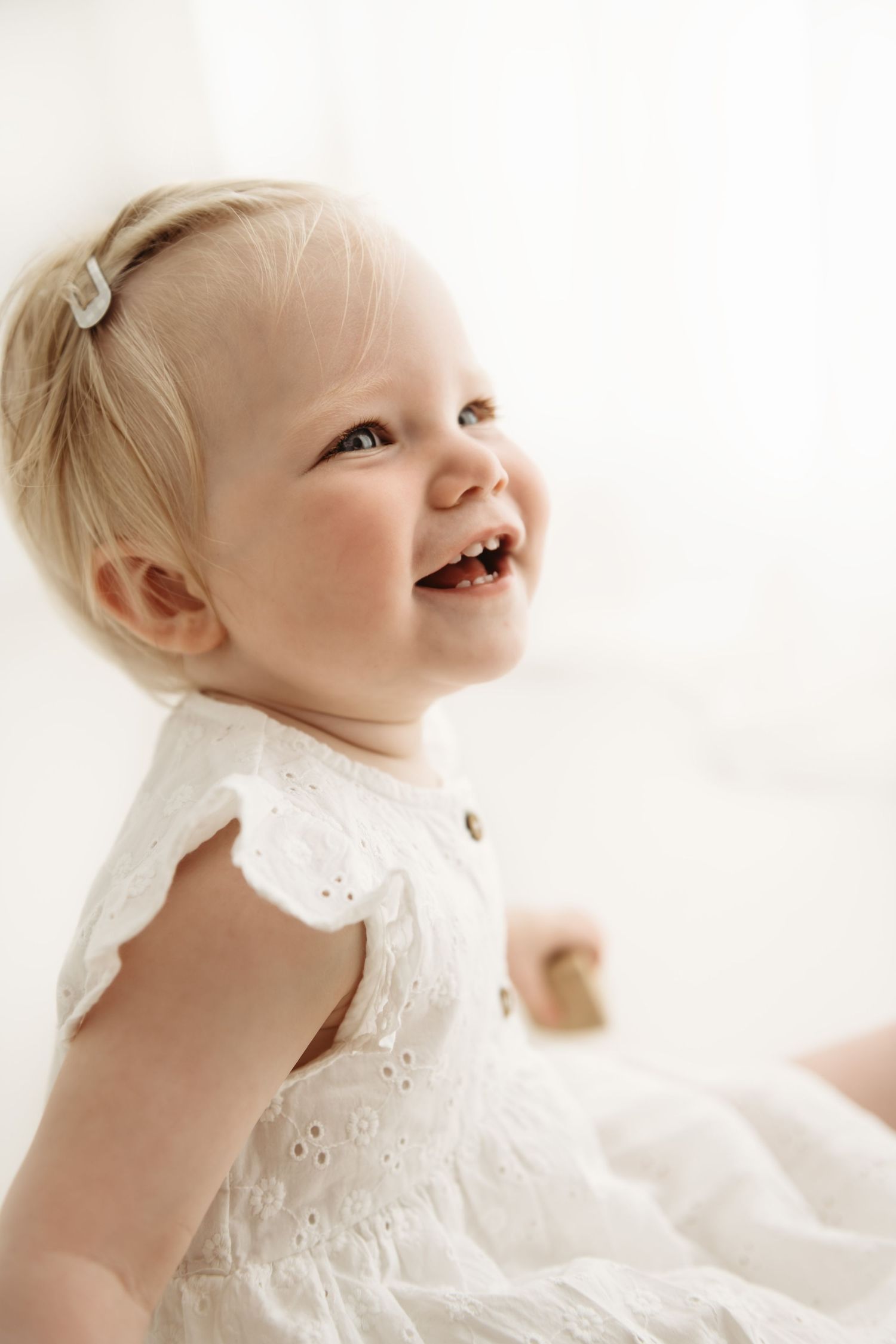 Baby wearing white eyelet dress smiles joyfully against white background.