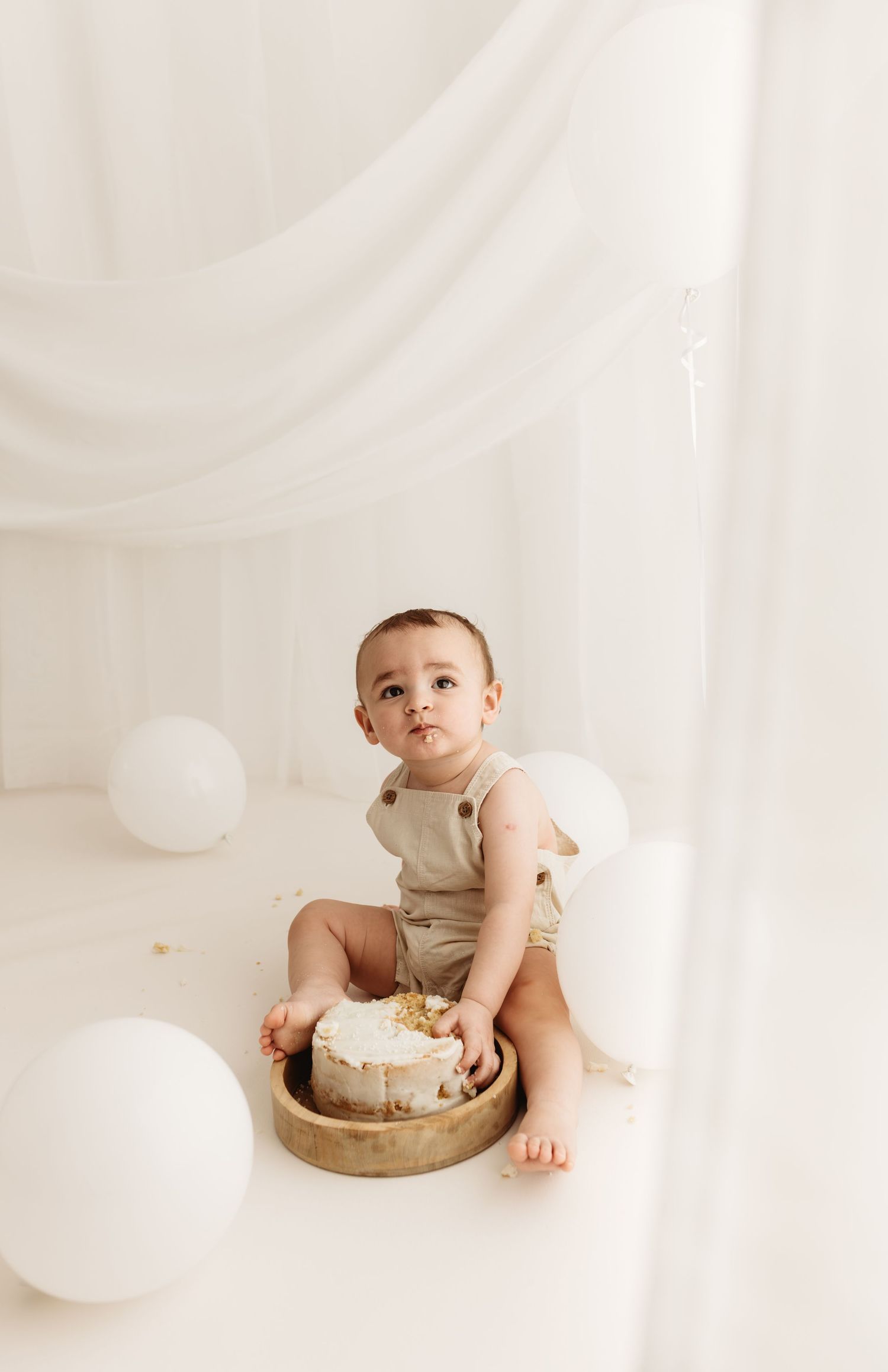 Baby in beige romper sits beside a smash cake surrounded by white balloons during a first birthday photo session.