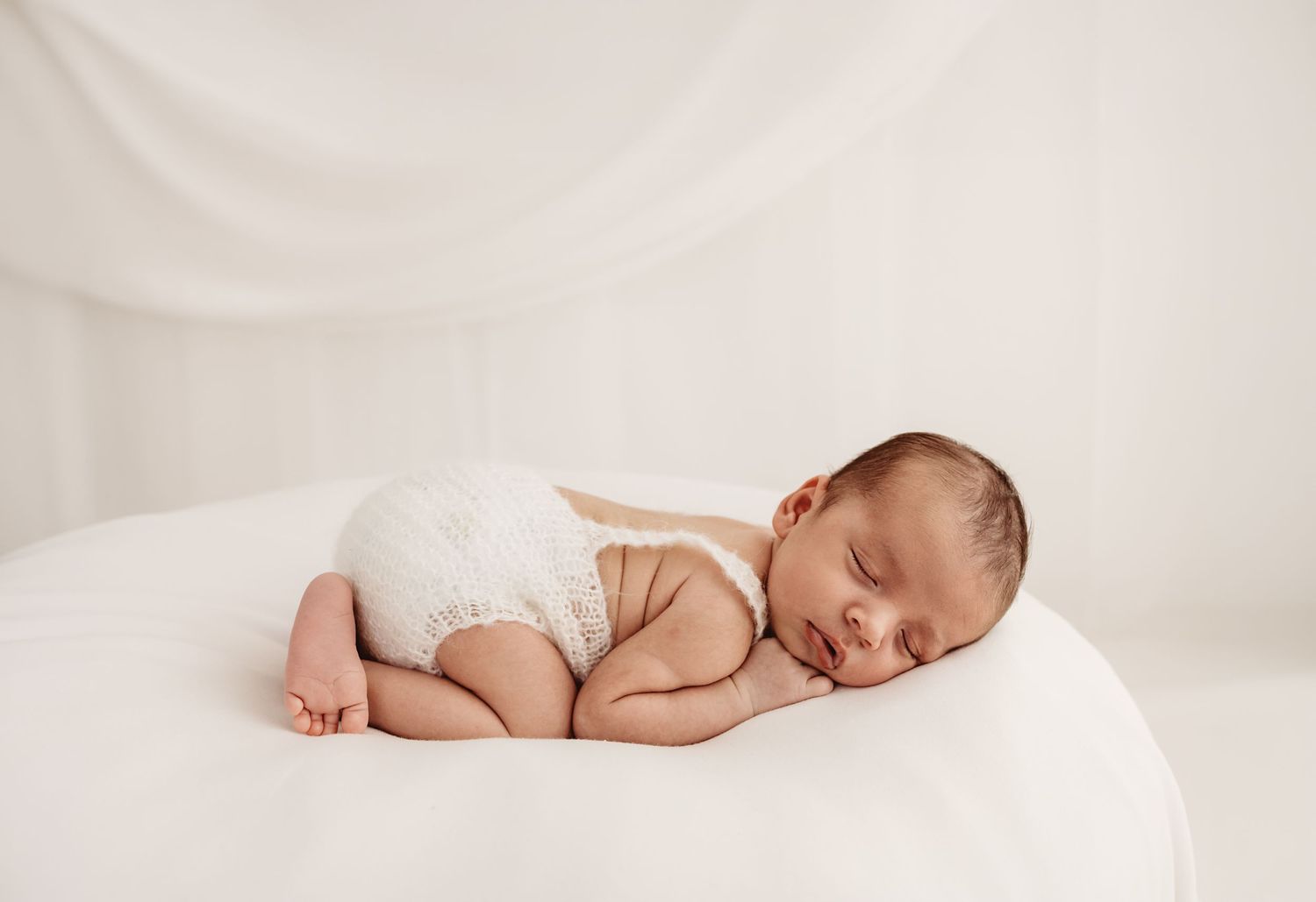 Peaceful newborn baby sleeping curled up on a white rounded surface, wearing a cream knit romper in a soft white studio.
