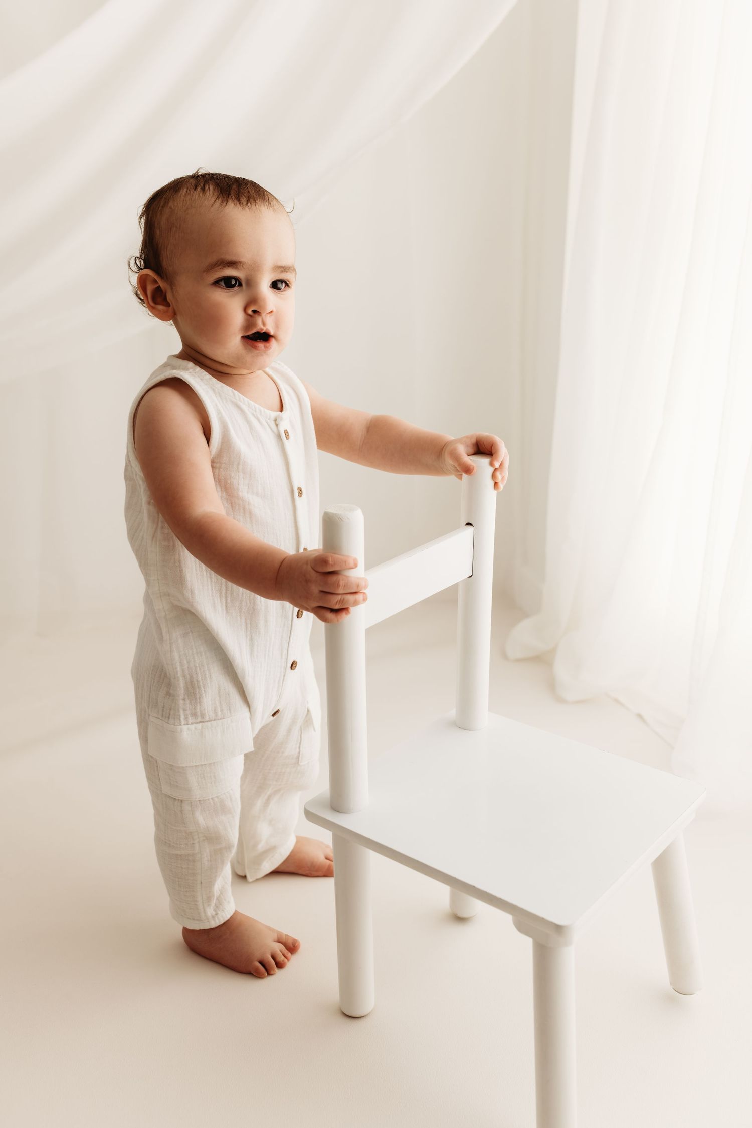 A baby in a white linen romper stands barefoot, holding a small white chair in a bright white studio setting.