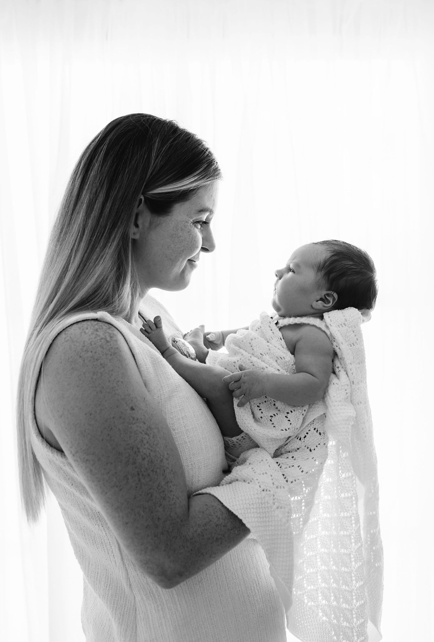 Black and white photo of a smiling mother tenderly holding her newborn baby, sharing a gentle moment by a bright window.