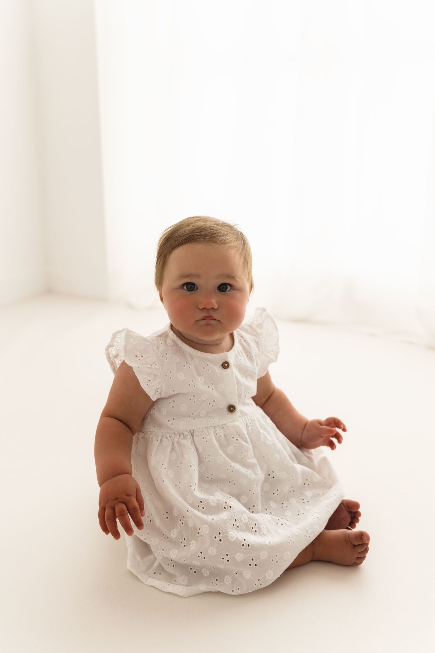 A baby in a white eyelet lace dress sits on a white seamless background in three similar poses.