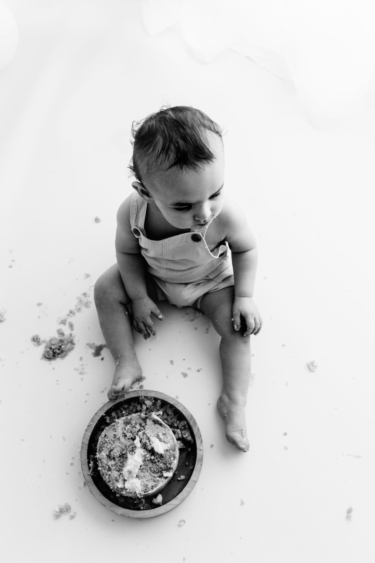 Black and white photo of a baby in overalls sitting on floor with a smashed first birthday cake.