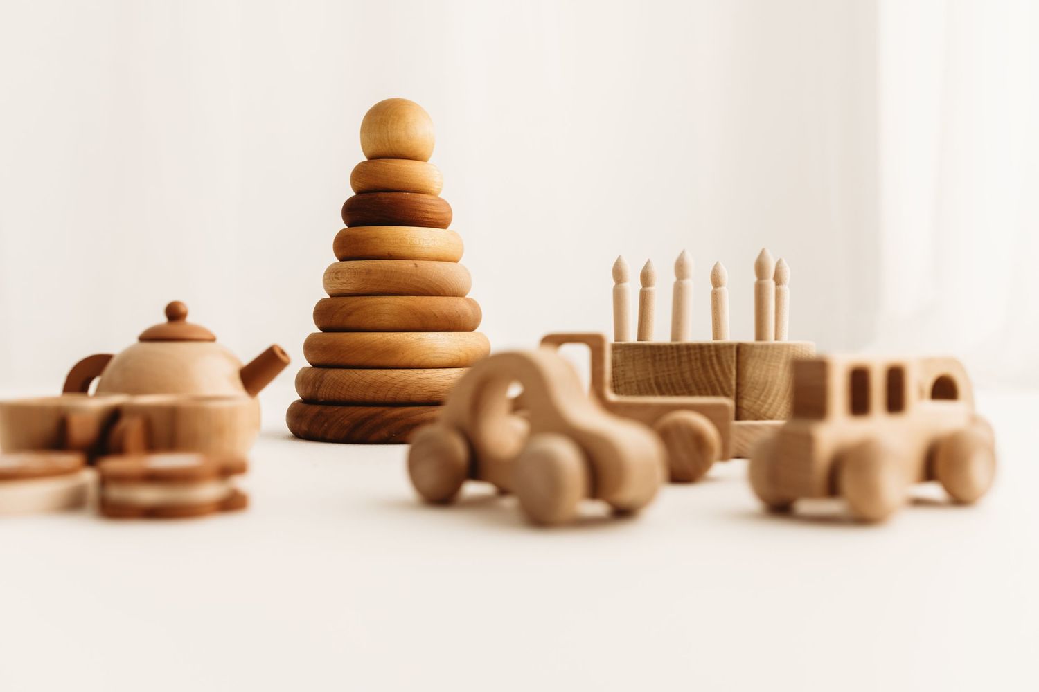 Natural wooden toys including a stacking ring pyramid, toy cars, birthday cake, and tea set arranged on a white surface.