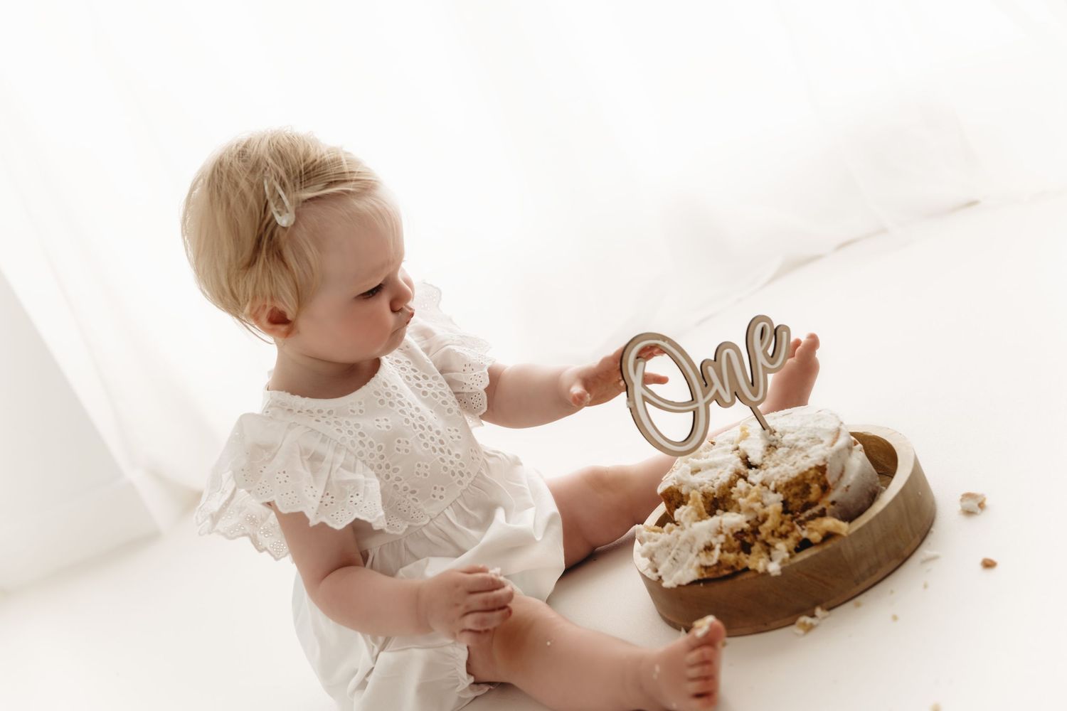 A sequence of four photos showing a baby in a white outfit smashing and eating cake during a first birthday celebration.
