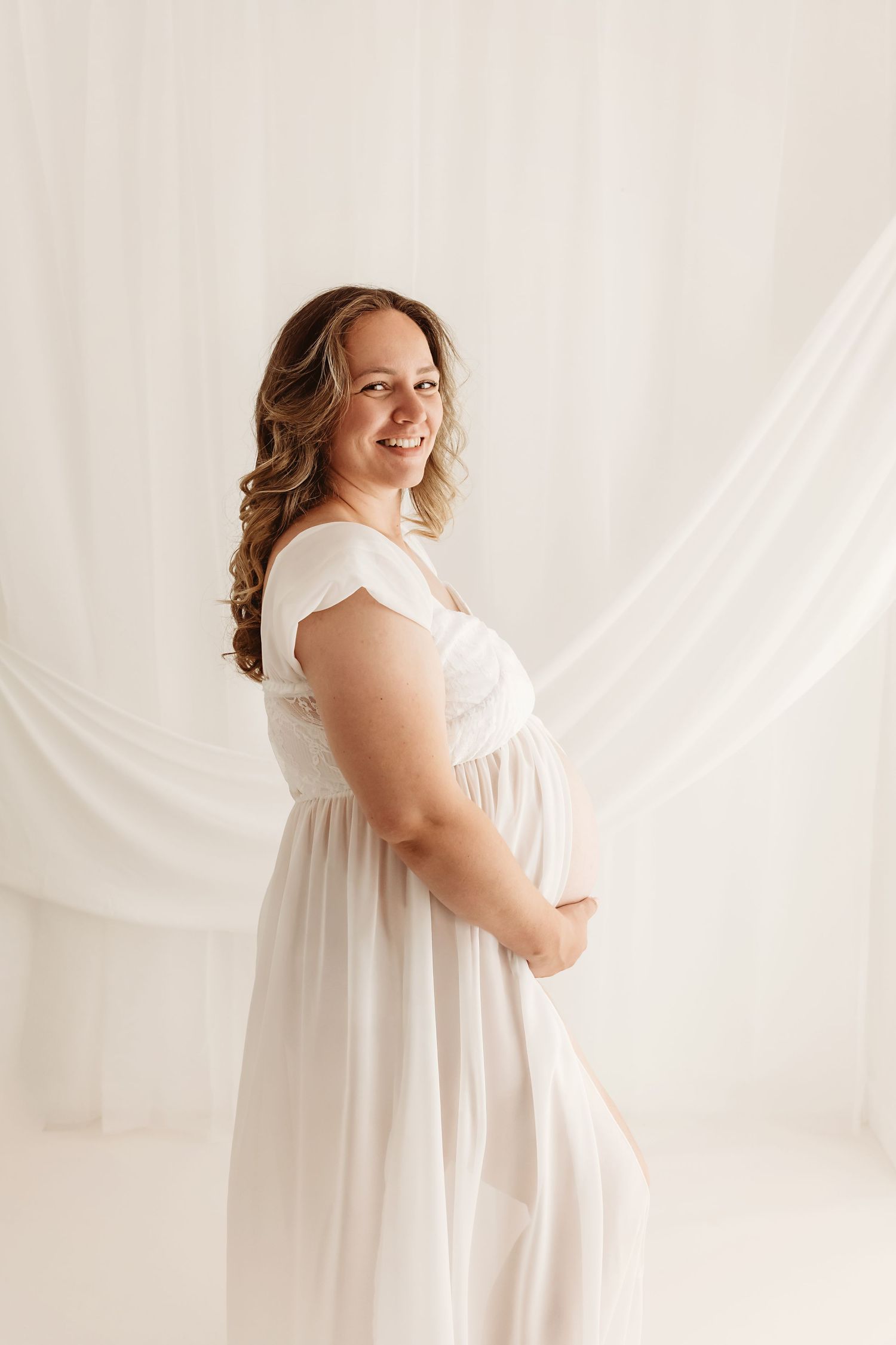 Smiling pregnant woman in white maternity gown poses gracefully against soft white draped fabric backdrop.