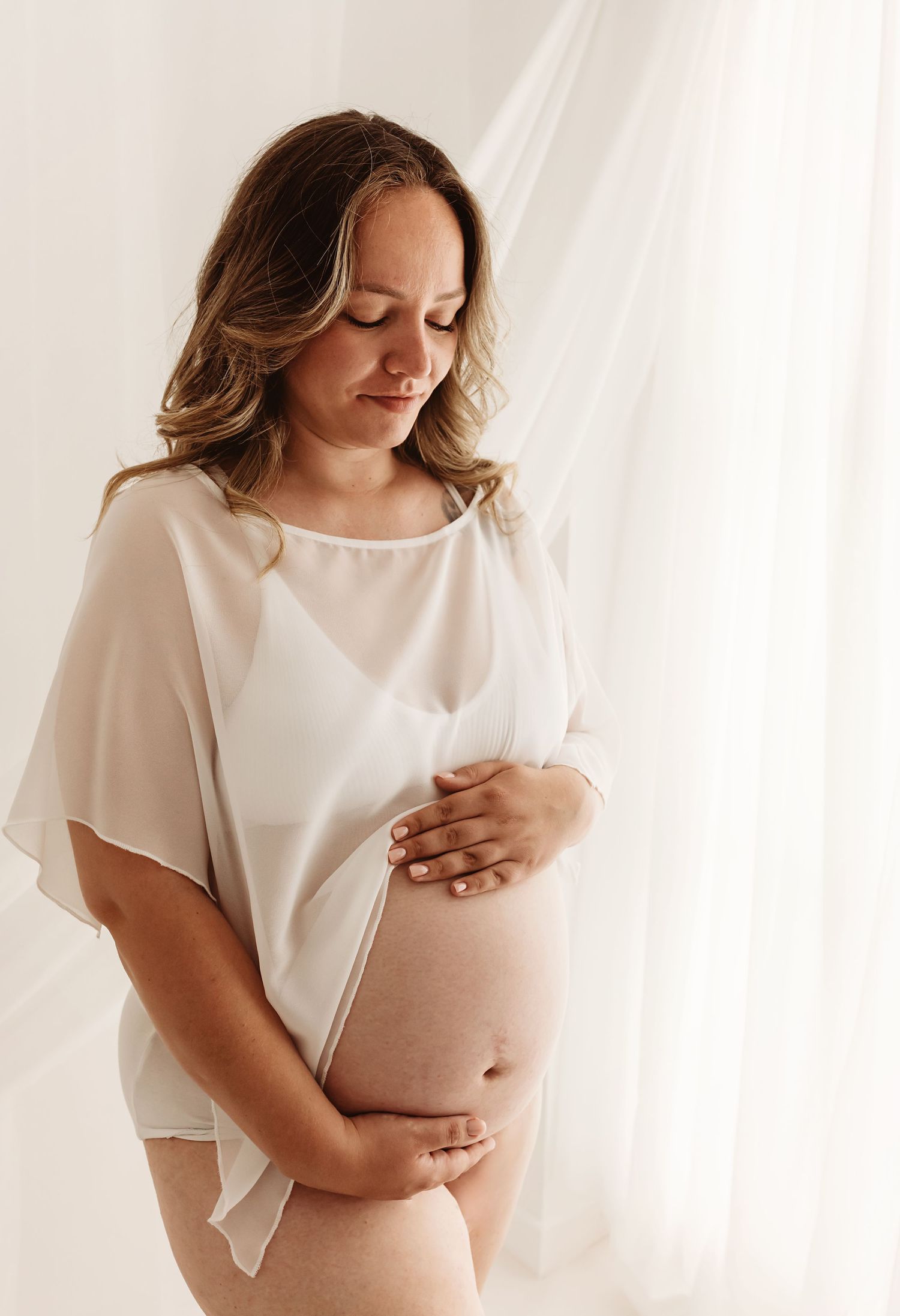 Pregnant woman in white top cradling her baby bump, standing by sheer curtains in a serene maternity photo.