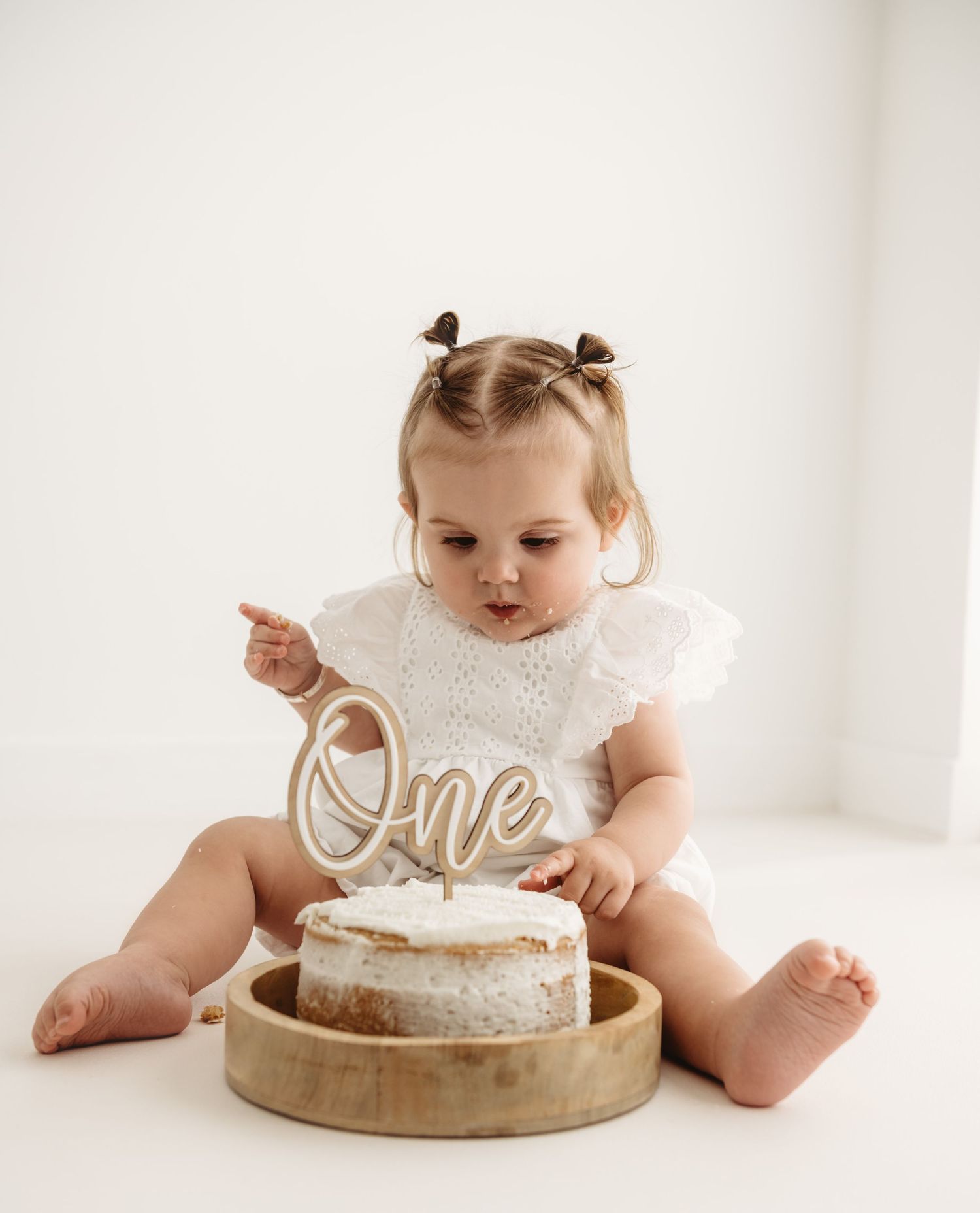 Baby girl in white romper touching her first birthday cake with 'One' topper during cake smash photo session.