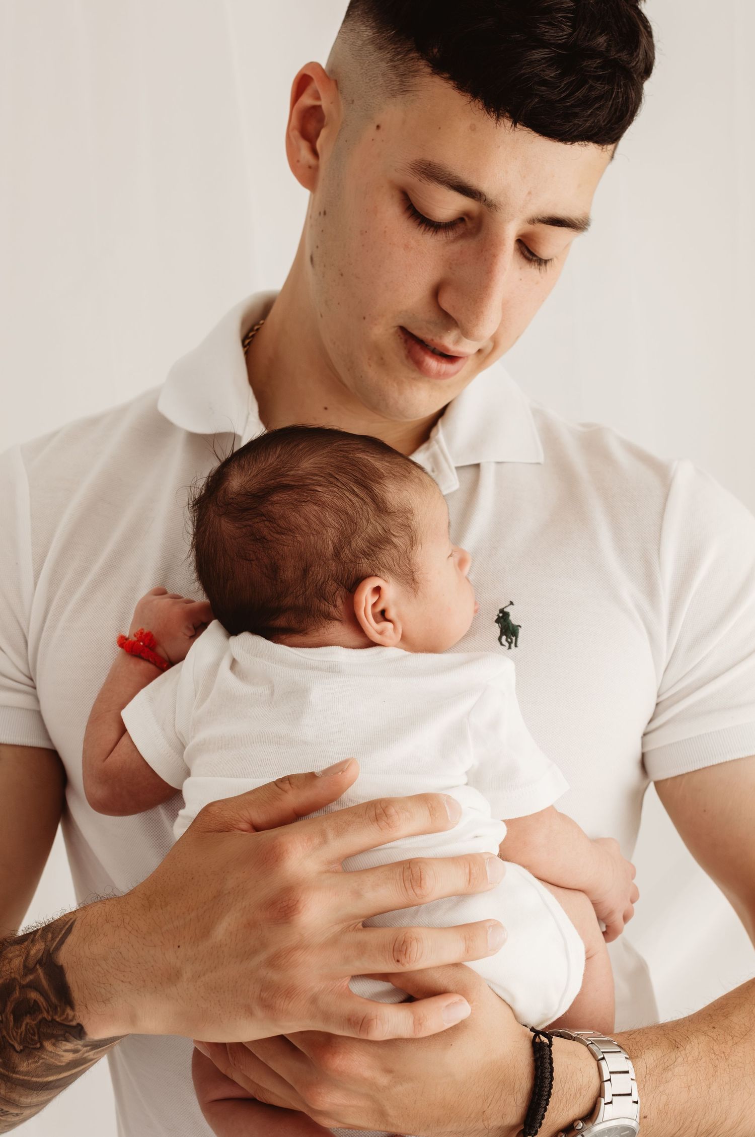 Young man in white Ralph Lauren polo shirt tenderly holding a newborn baby in white onesie with red hospital bracelet.