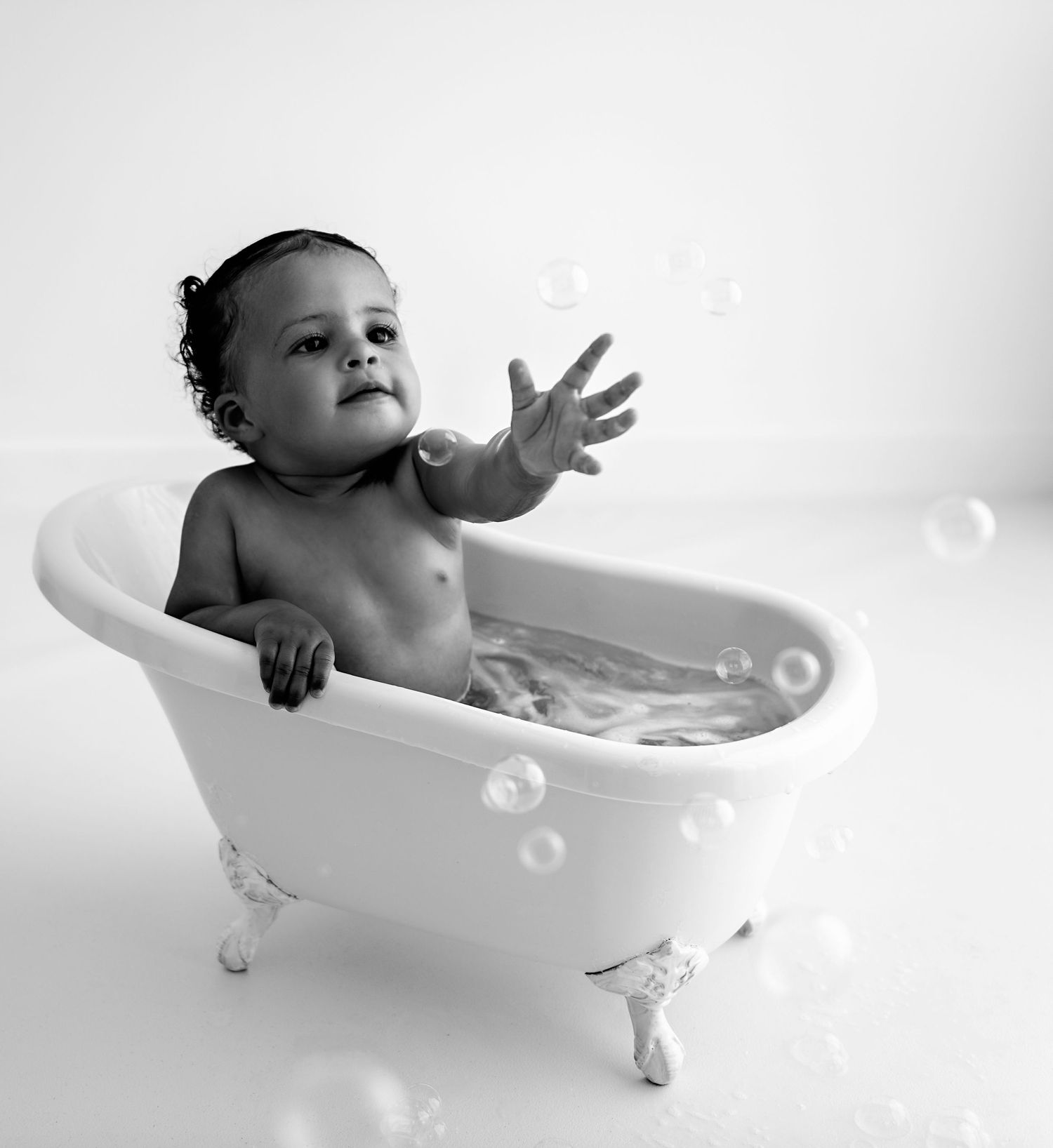 A baby plays with bubbles and splashes in a white vintage bathtub during bath time in black and white photo series.
