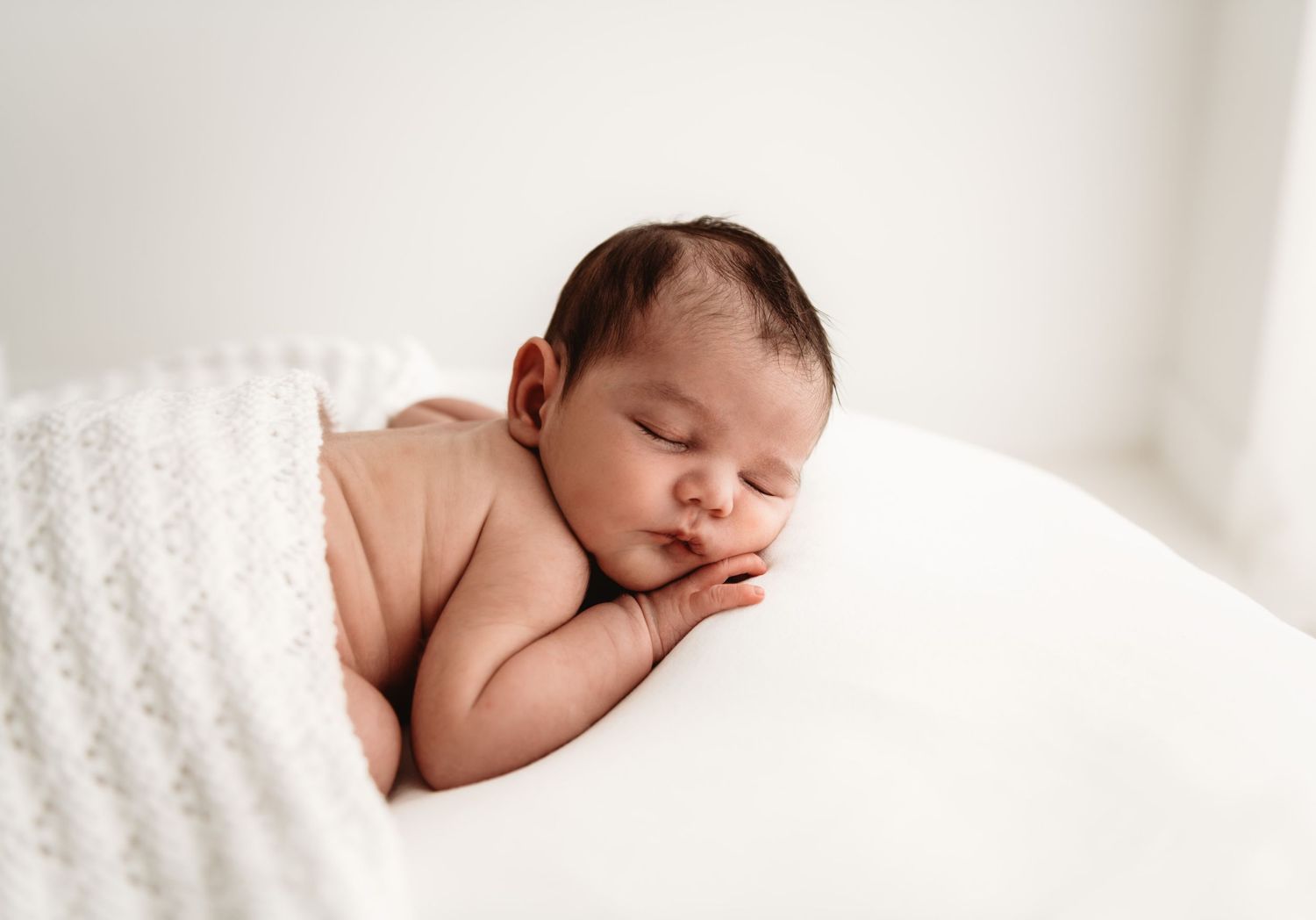 Peaceful newborn baby sleeping curled up on white surface with soft blanket, professional newborn photography.