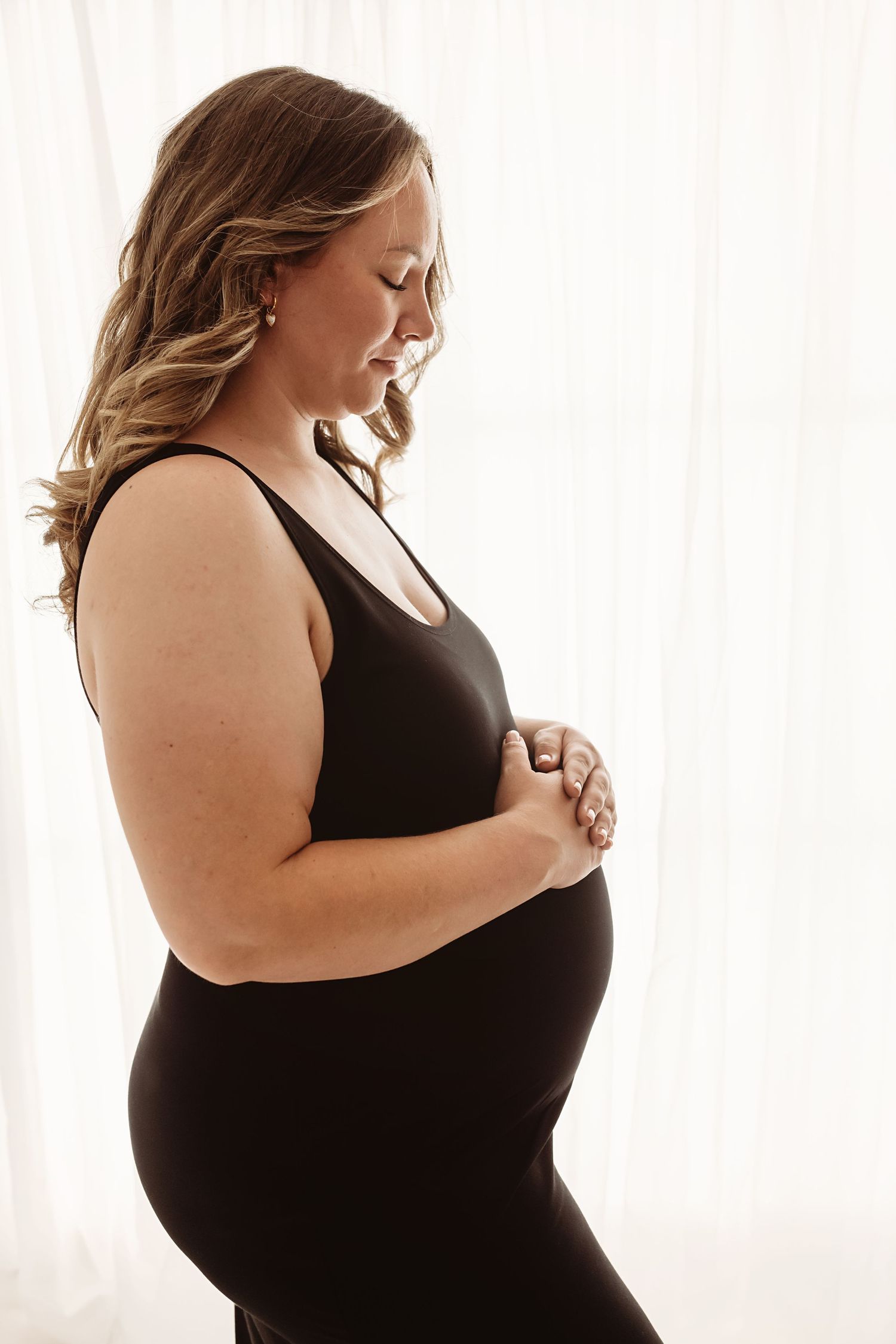 Pregnant woman in black dress cradling baby bump, standing in soft natural light against white curtains.