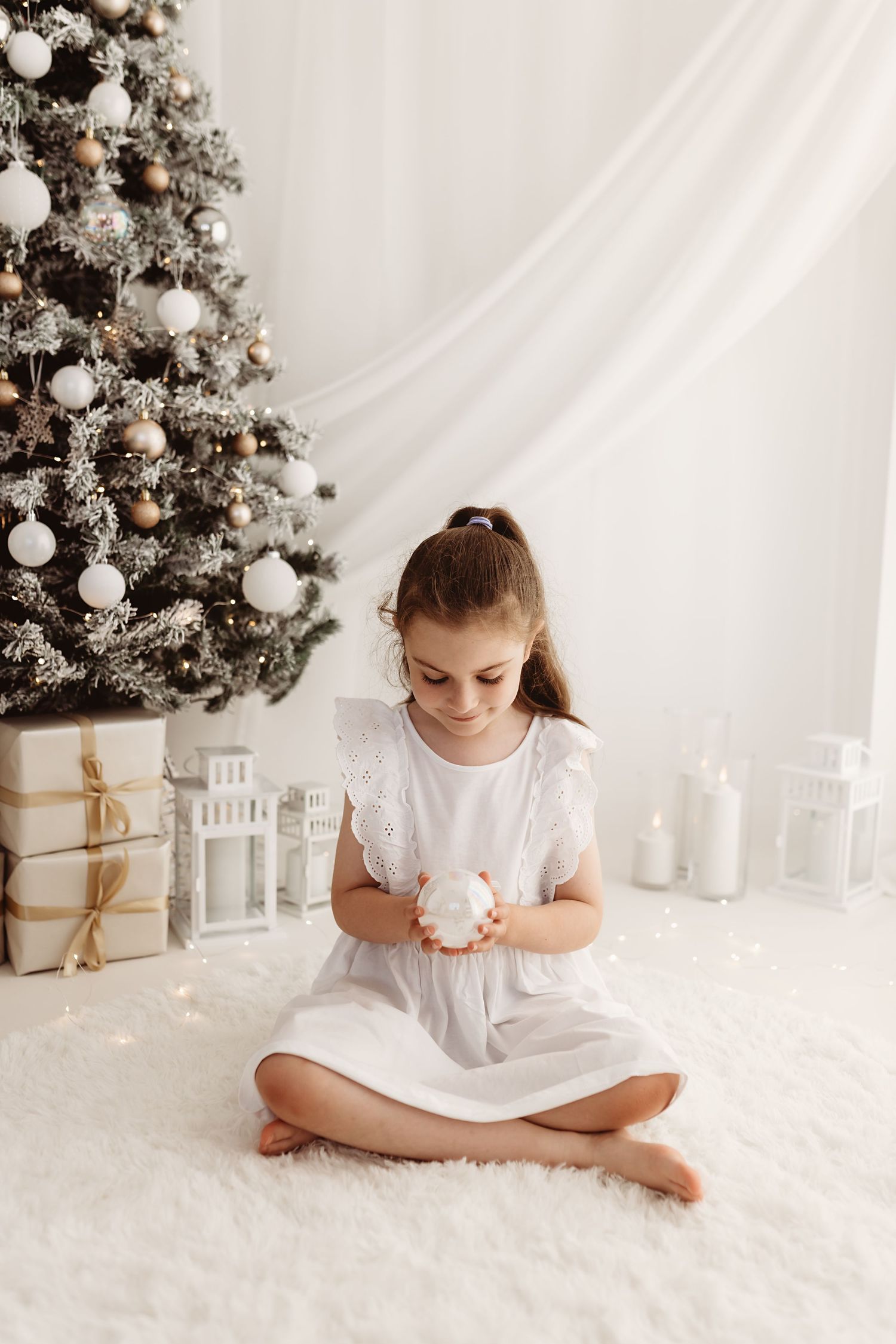 A young girl in a white dress sits on a fluffy rug holding a ornament near a decorated Christmas tree with gifts.