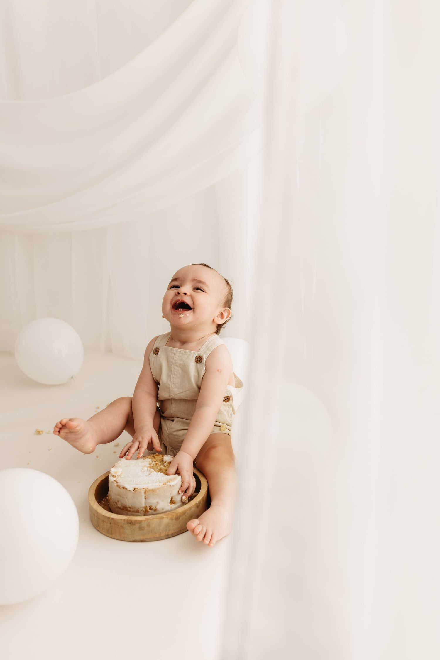 Laughing baby in beige romper smashing first birthday cake in white studio with balloons.