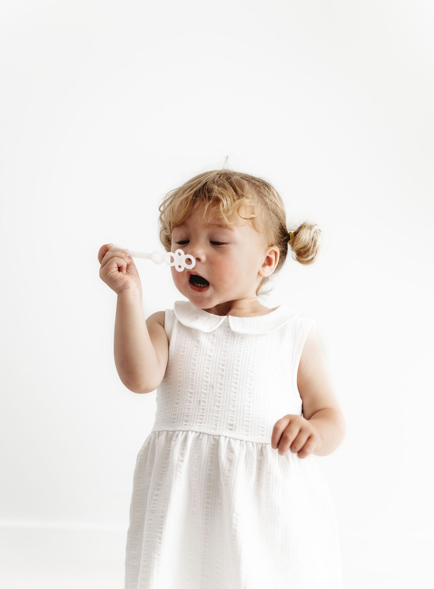 Toddler in white sleeveless dress holds small bubble wand against light background.