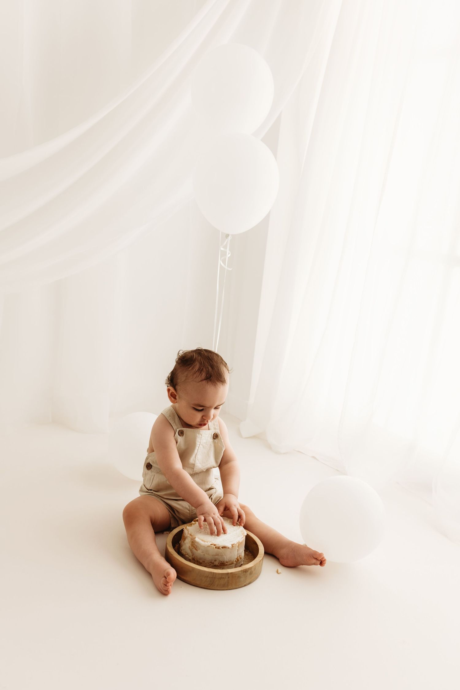 Baby in beige romper smashing first birthday cake on white floor with white balloons and sheer curtains during a cake smash session in Corby
