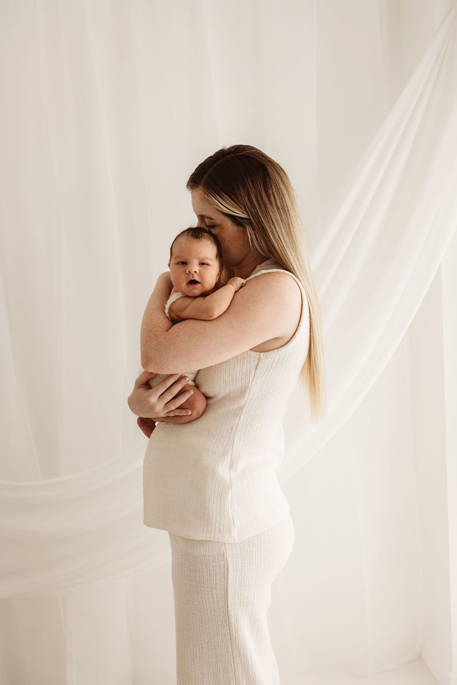A mother in a white outfit tenderly holds her newborn baby against a soft white draped background.