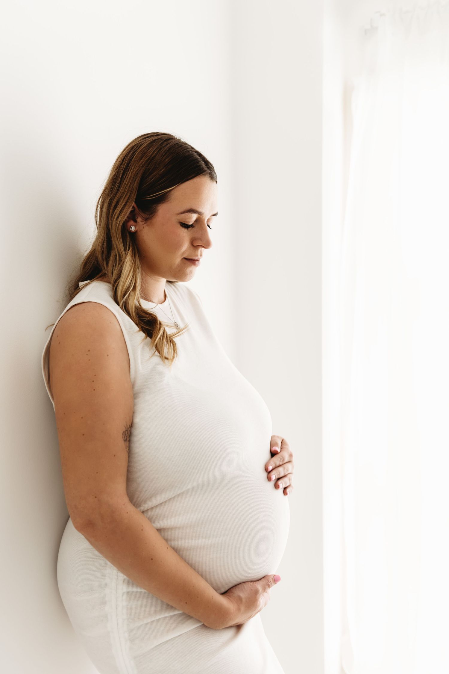 Pregnant woman in white dress cradling her baby bump against a white wall in a serene maternity photo.