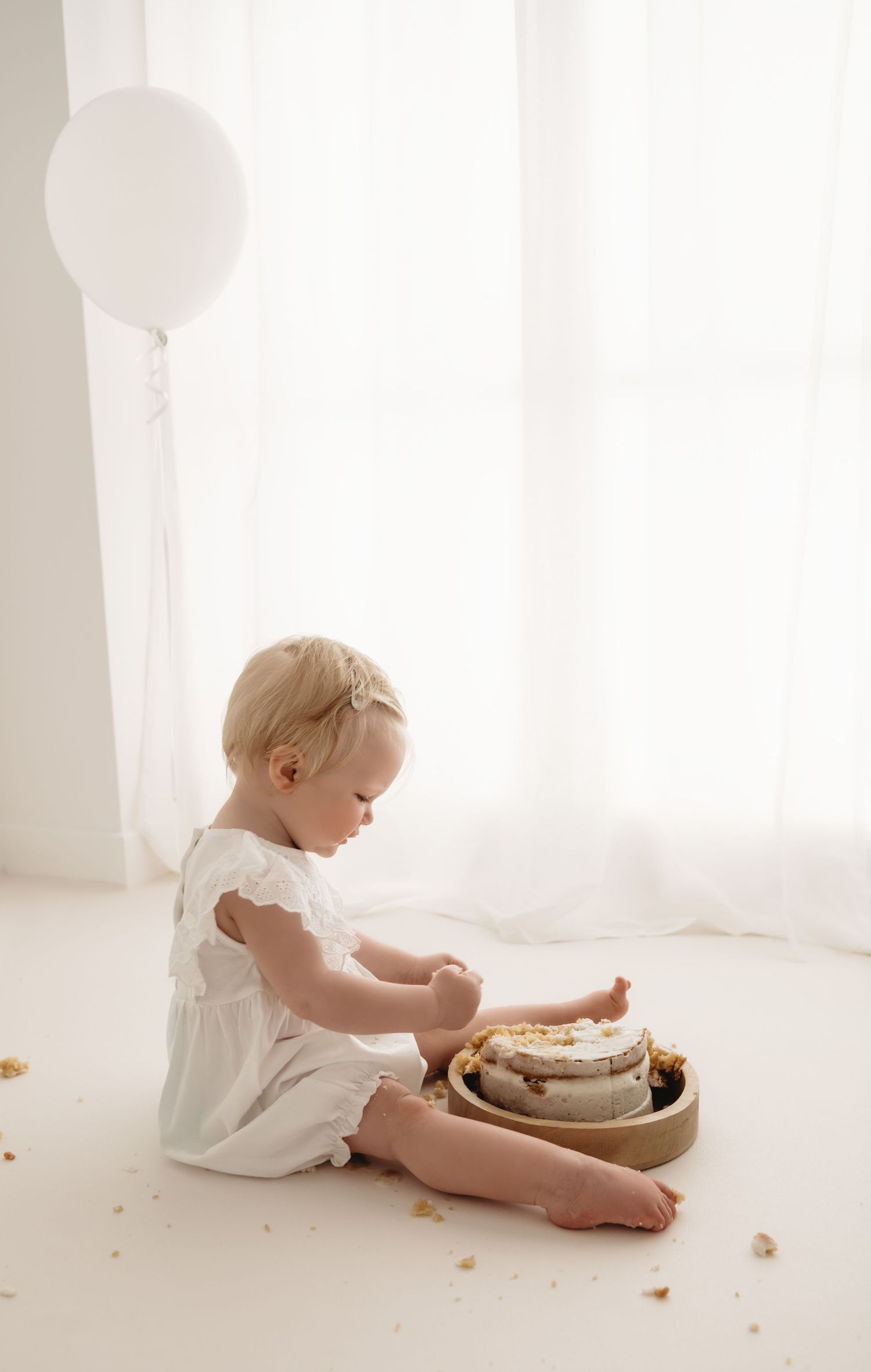 A baby sitting side on with a cake in-between her legs and a birthday balloon during a Cake smash session in Corby, Northamptonshire.