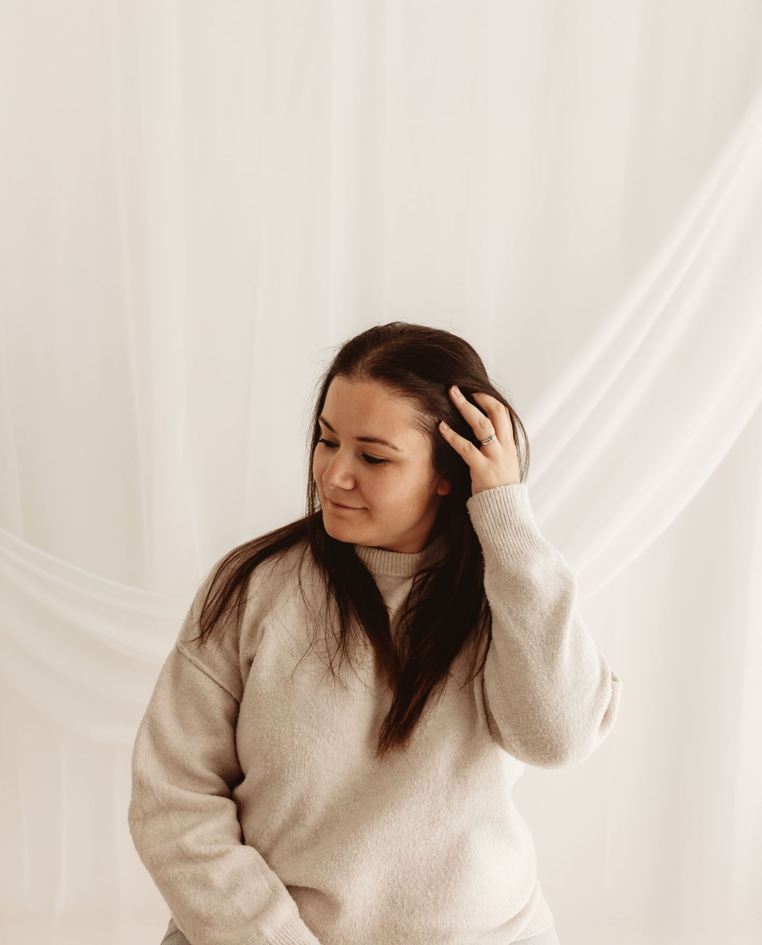 Young woman in a beige sweater poses with hand in dark hair against a soft white curtain backdrop.
