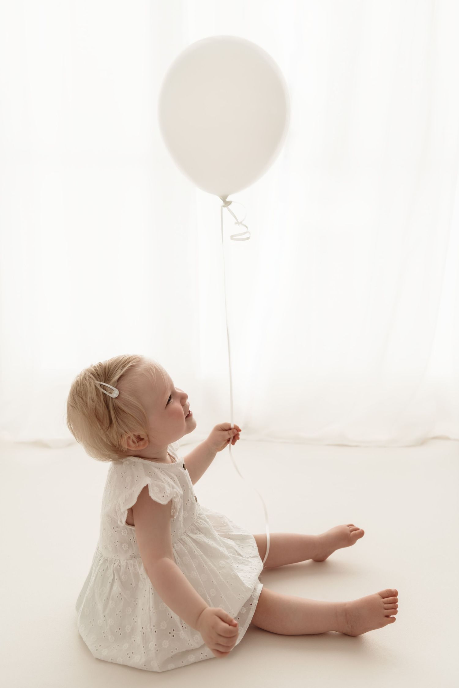 Blonde toddler girl in white eyelet dress sitting barefoot on floor holding a white balloon.