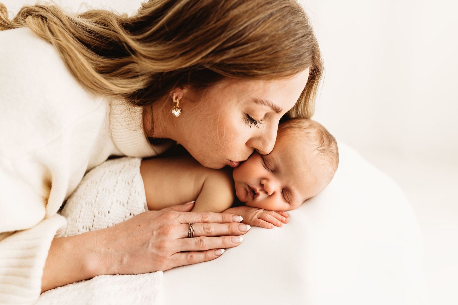 A tender mother gently kisses her sleeping newborn baby, both dressed in soft white, in a bright minimalist setting.