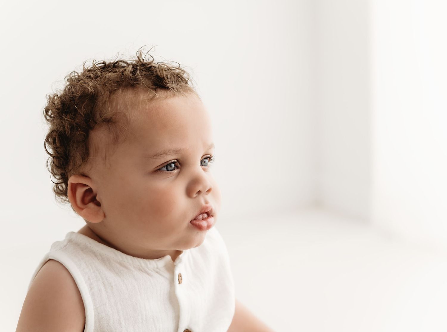 Baby wearing white clothing looking to the side against bright white background.