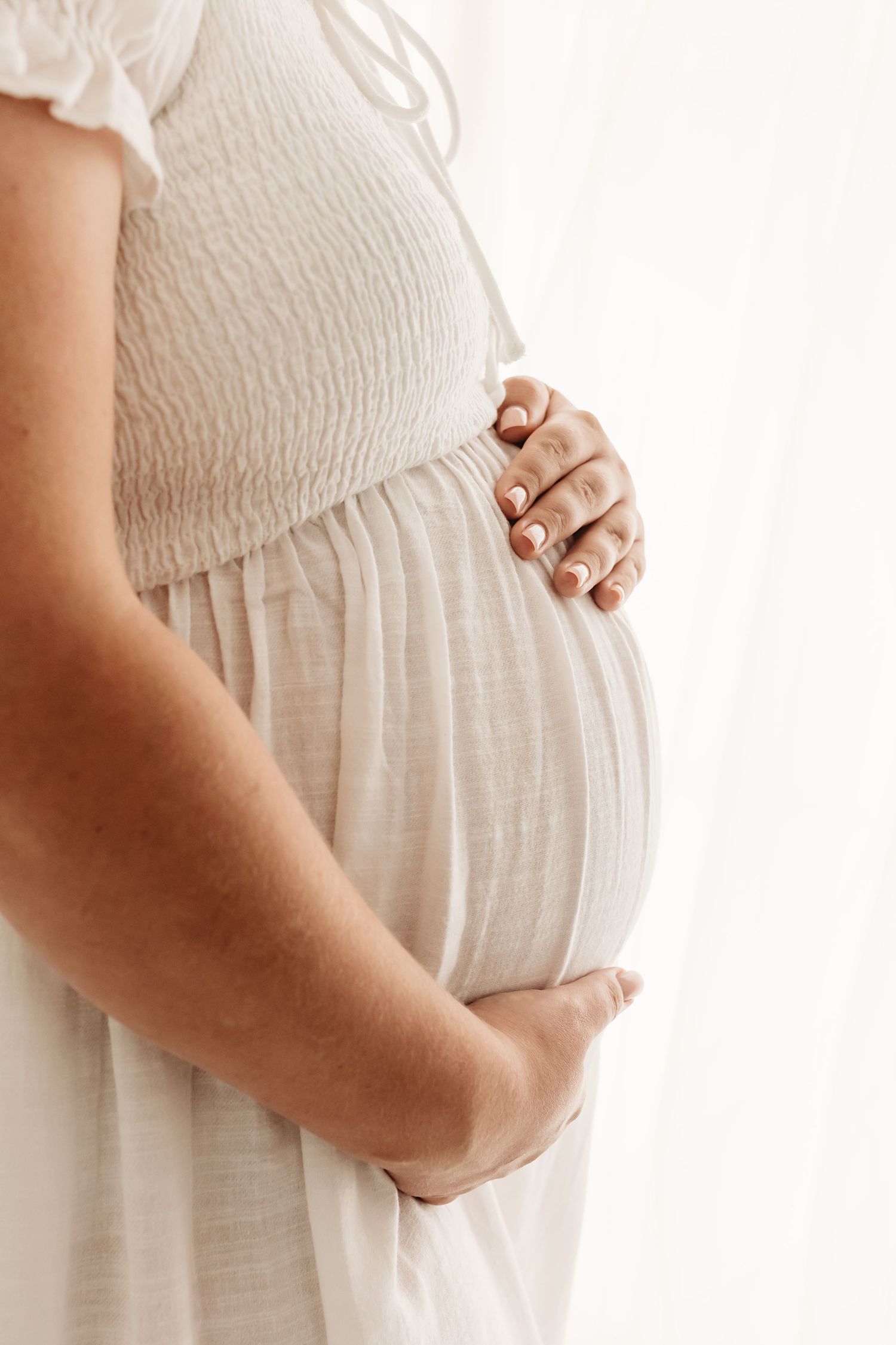 Pregnant woman in white dress cradling her baby bump with both hands against a soft white background.
