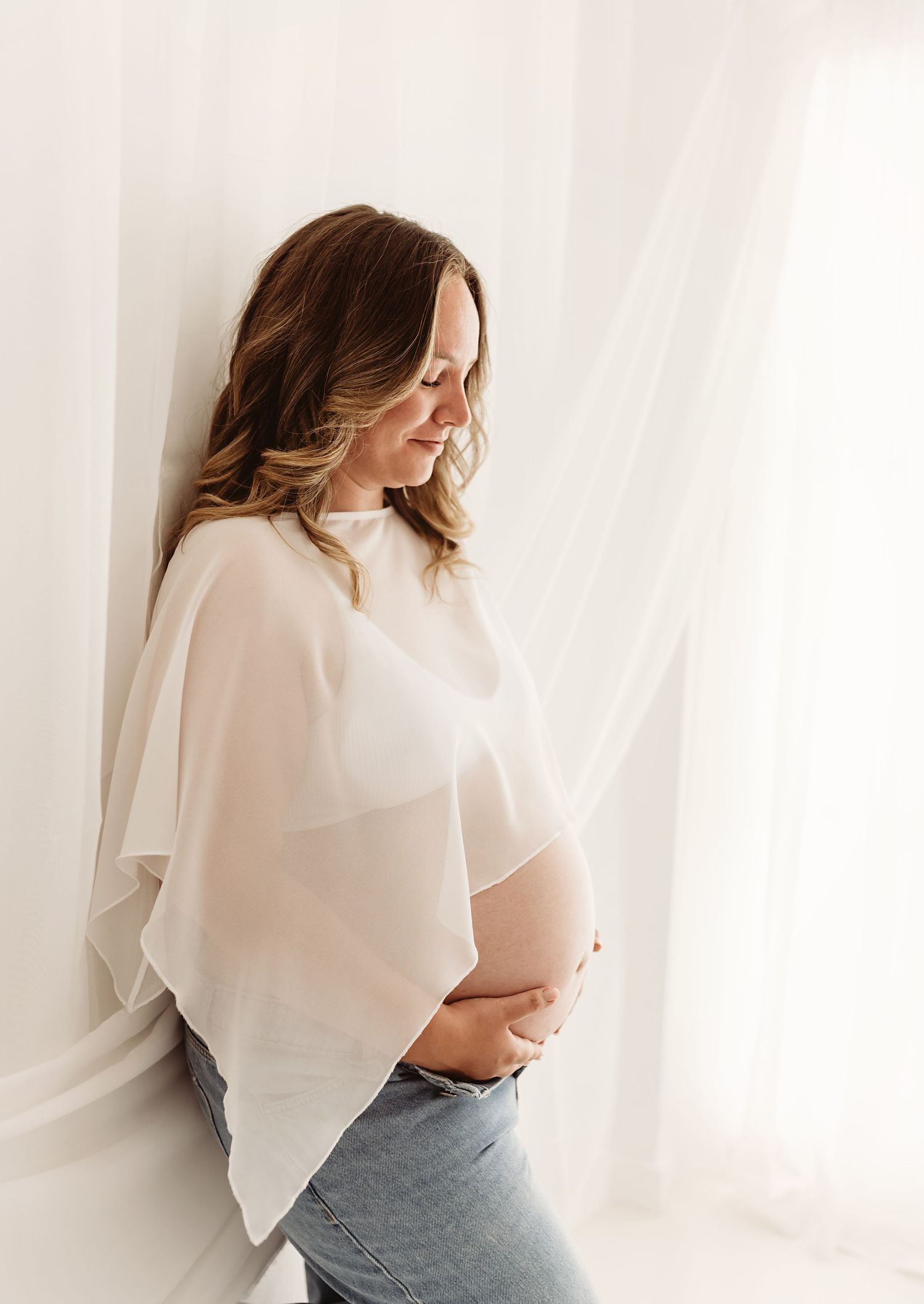 Pregnant woman in sheer white top and jeans cradling her baby bump, posing gracefully against soft white curtains.