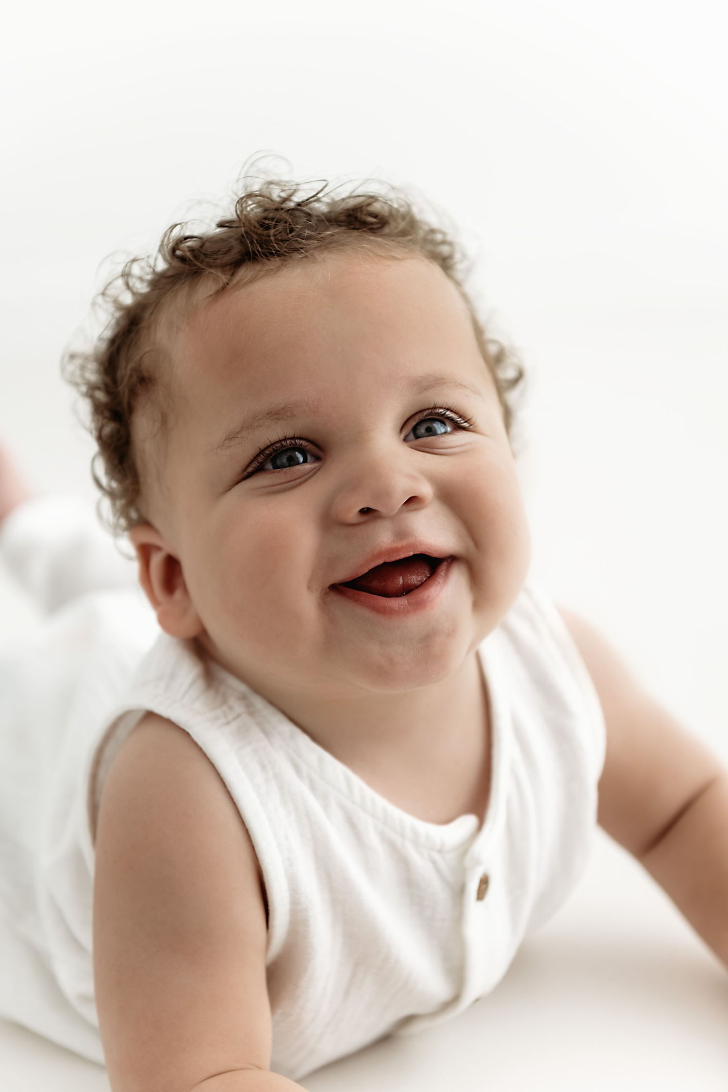 A happy baby with curly hair wearing a white sleeveless outfit smiles brightly while lying down on a light background.