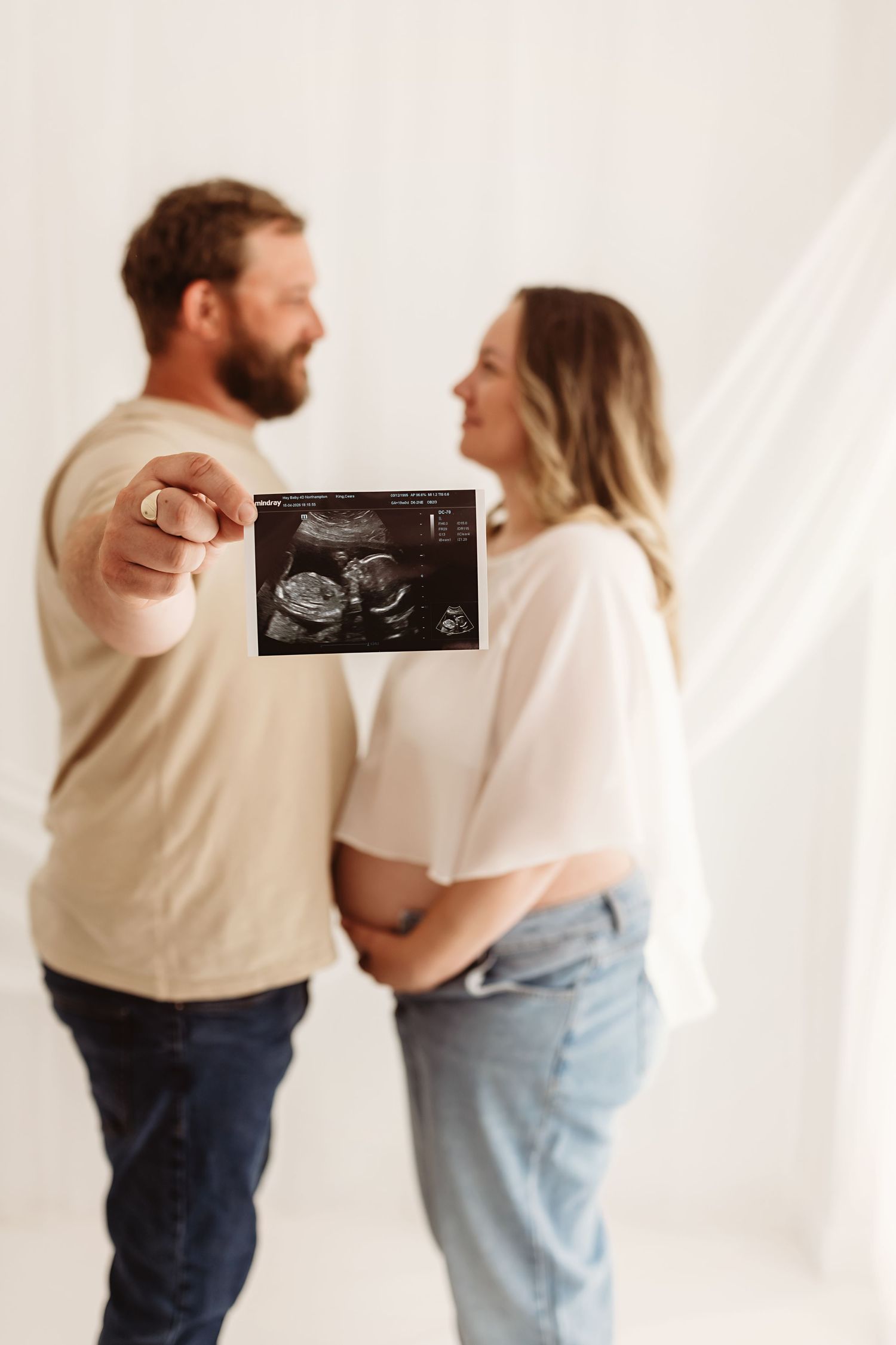Happy couple holding ultrasound scan photo, pregnant woman cradling baby bump in white studio setting.