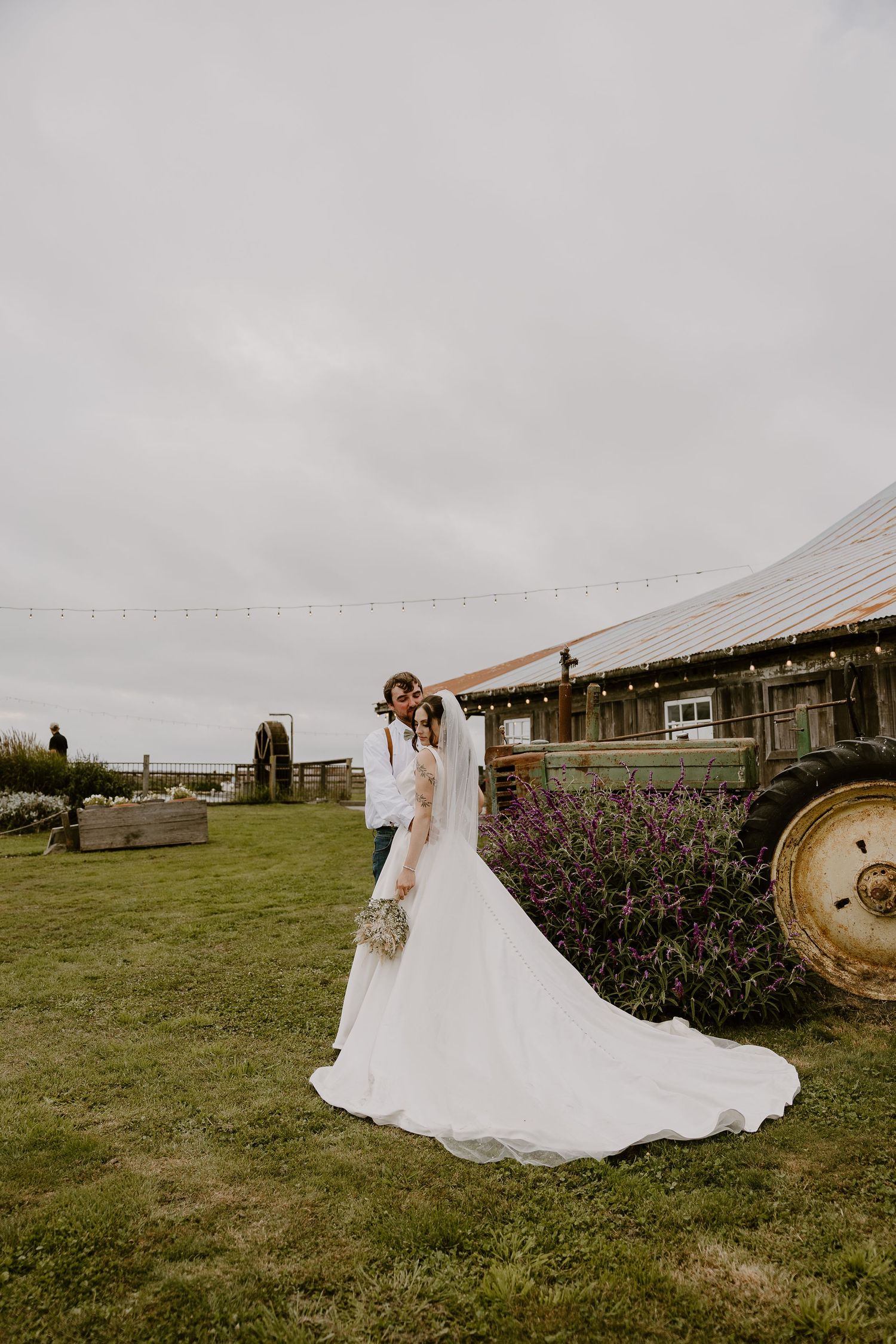 A rustic farm wedding scene with a white bridal gown against a weathered barn and vintage tractor on a cloudy day.