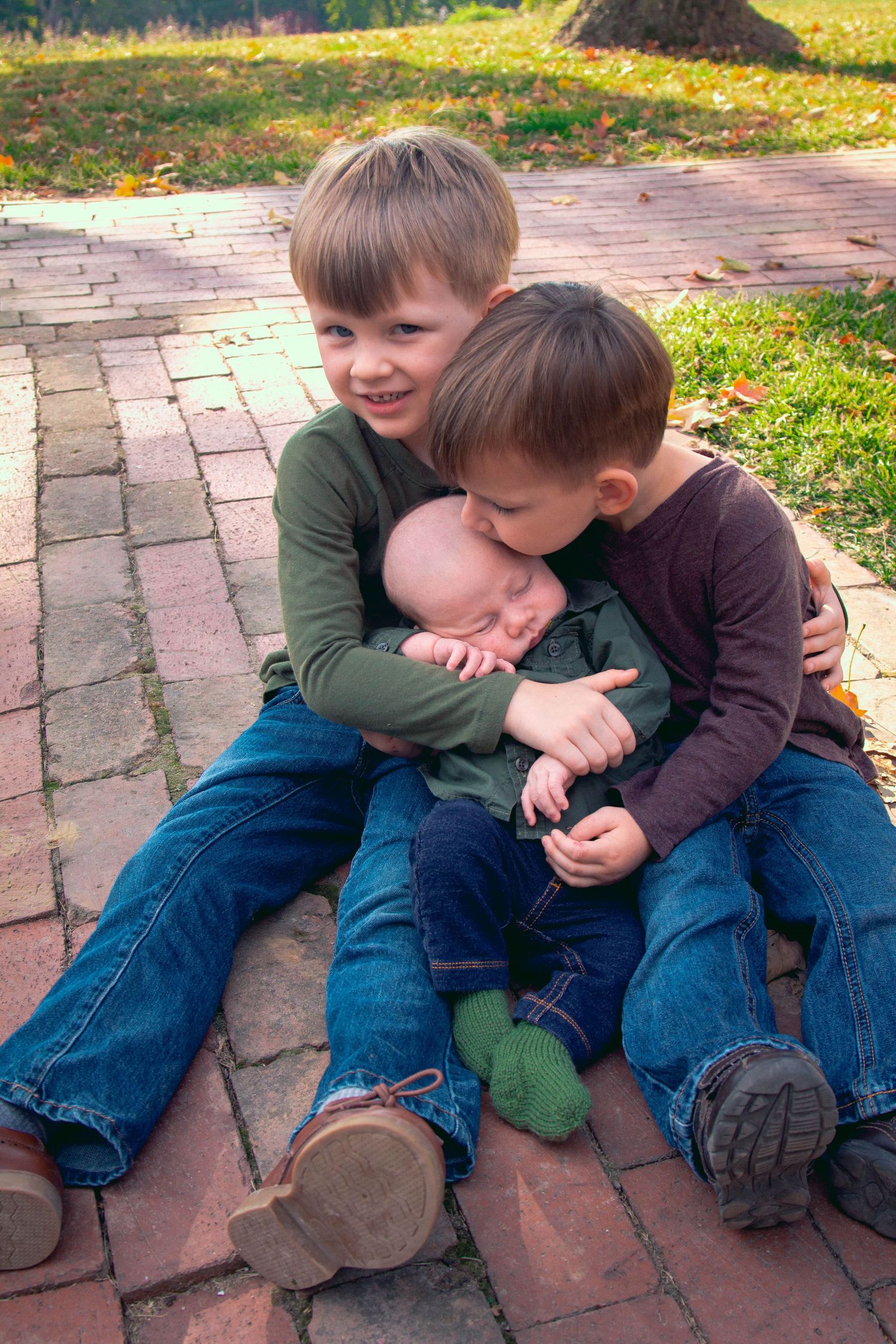 two older brothers are holding their baby brother and giving him a kiss on the head, they are wearing blue jeans and brown and green