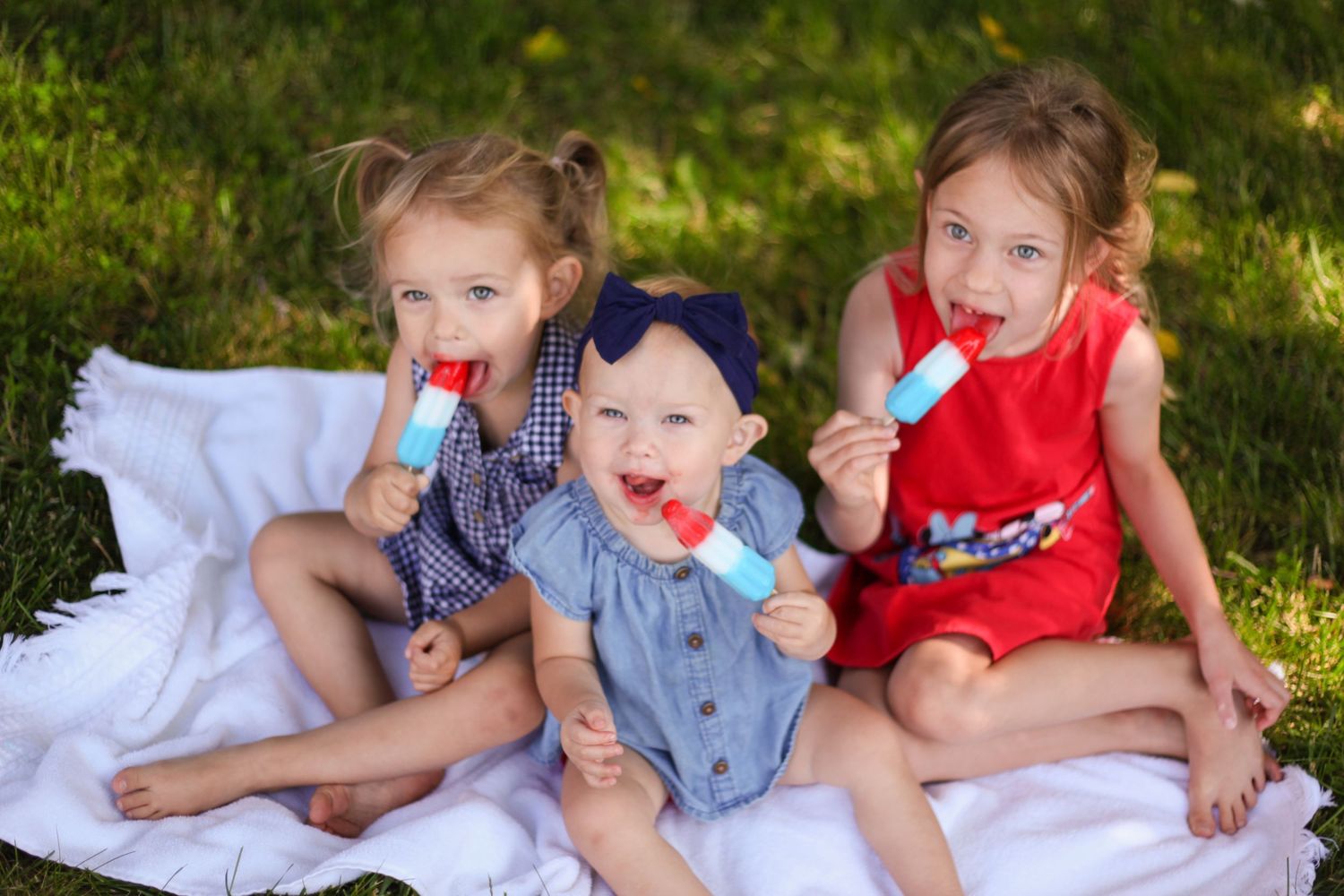 three girls are eating bomb popsicles on a white blanket wearing red, white and blue outfits and bows in their hair