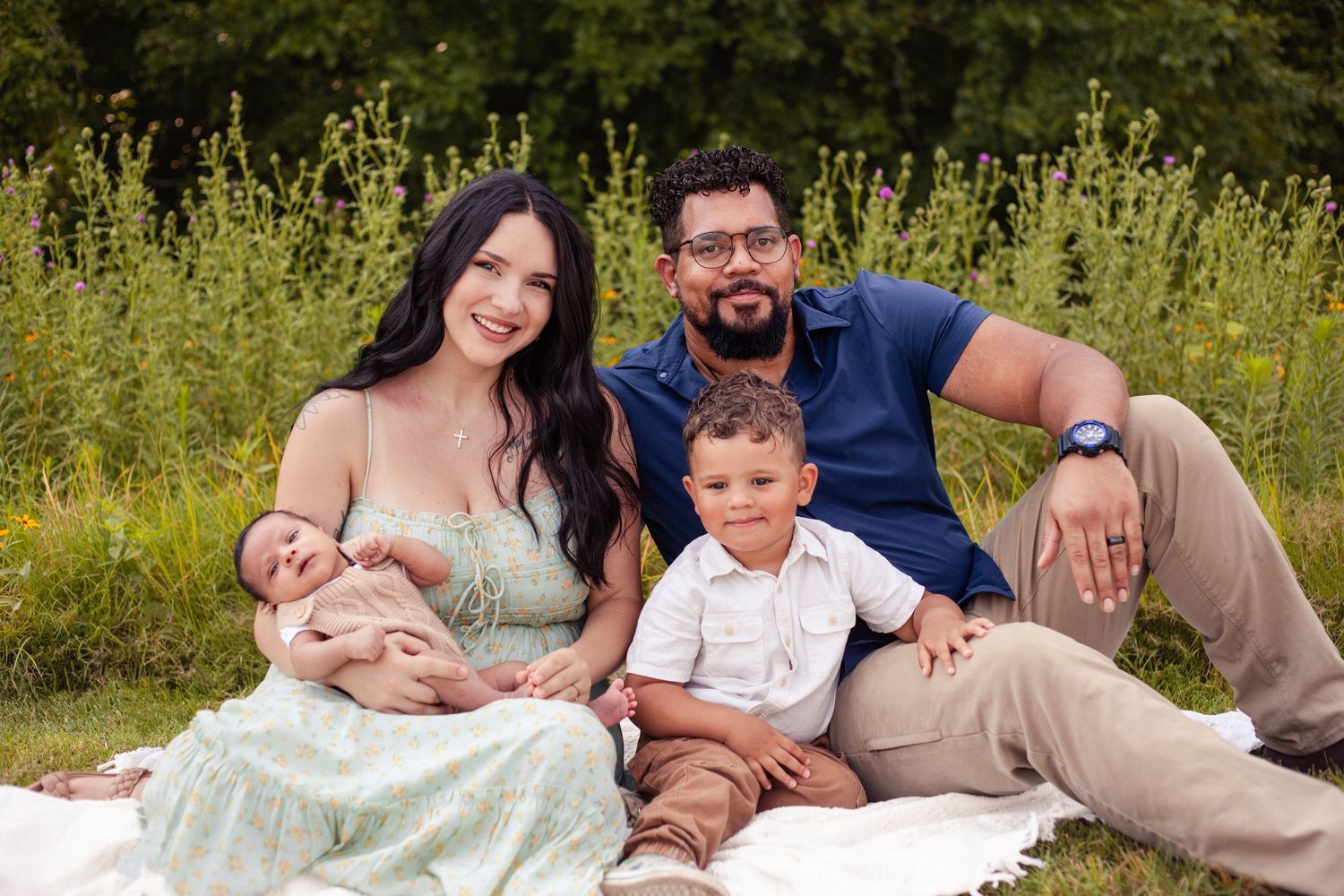The family is sitting with mom, dad, toddler boy and newborn boy. They are wearing green, white, blue and beige with flowers behind them.